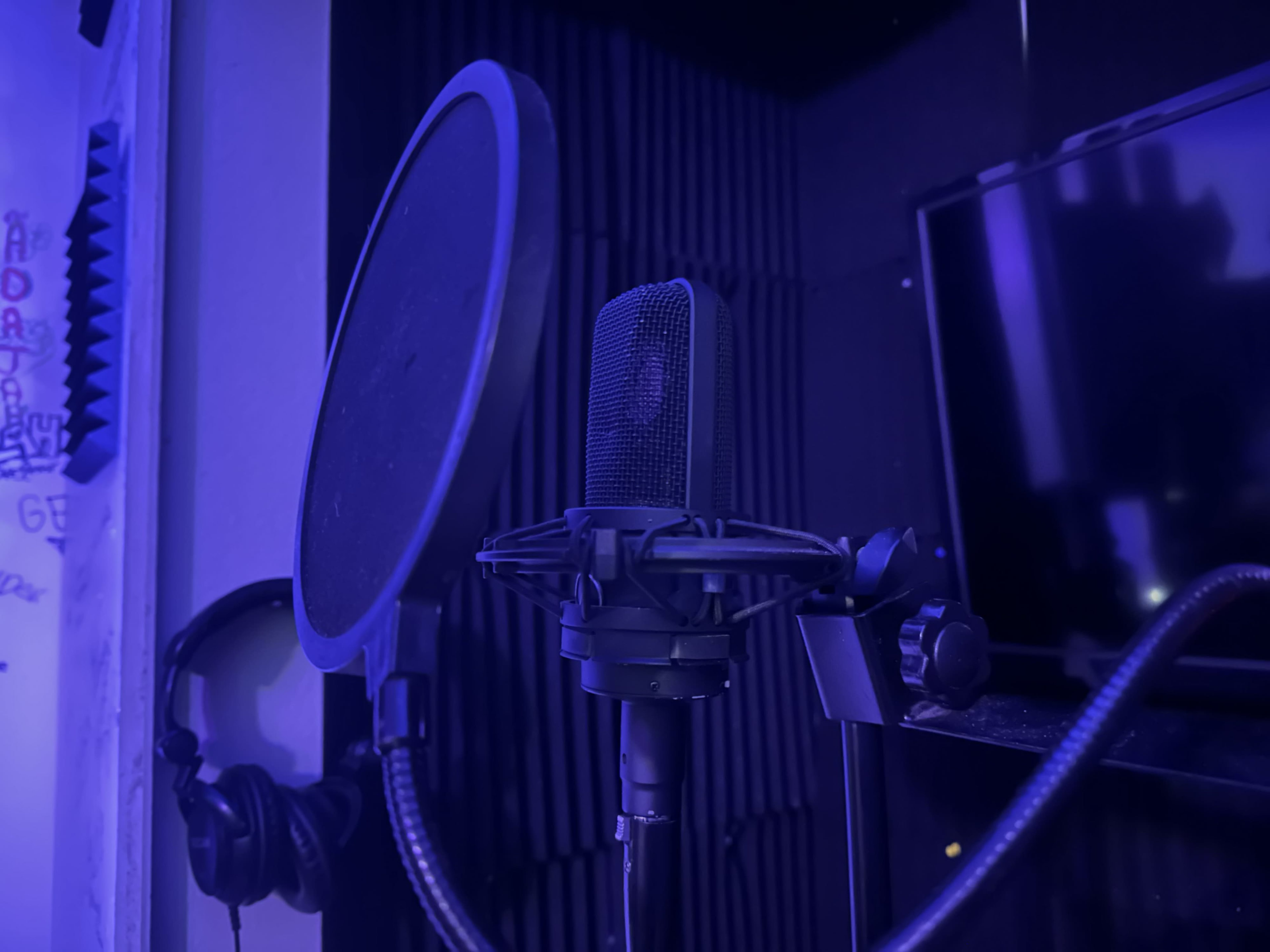 A microphone with a pop filter is positioned in a sound booth illuminated by blue light, with headphones hanging nearby.