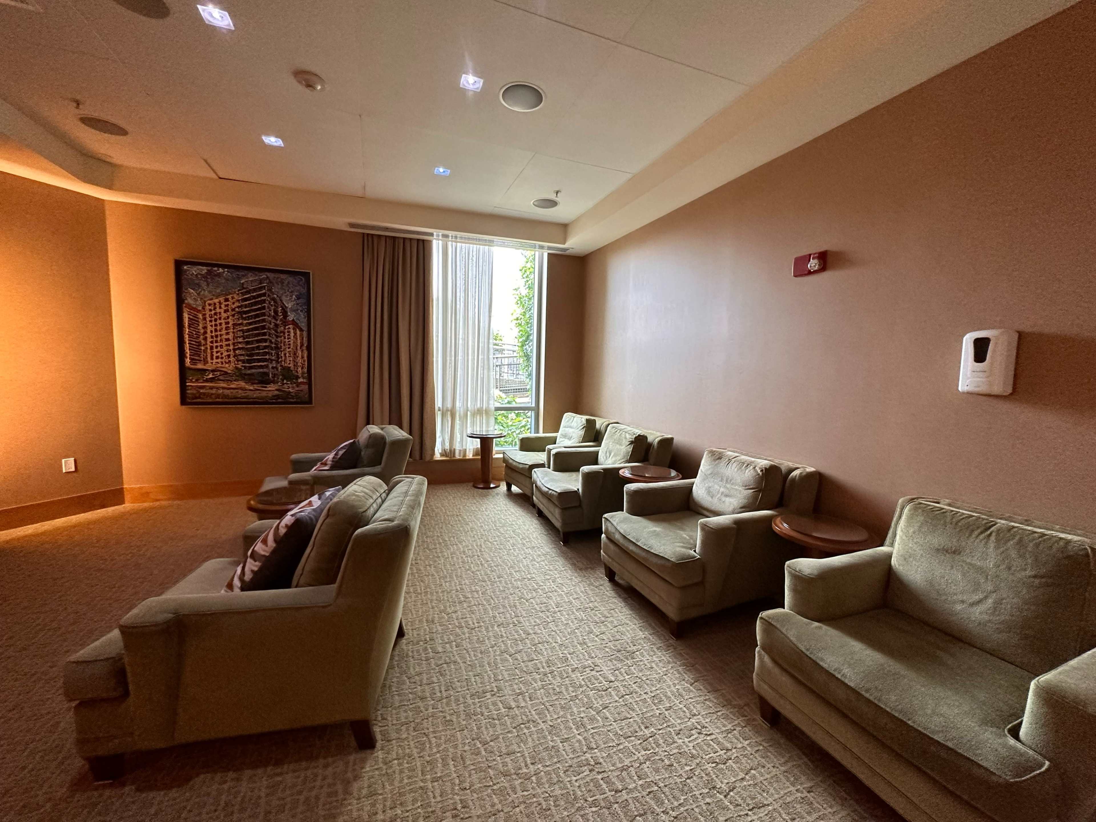 The image shows a small lounge area with green upholstered chairs, a side table, and a large window allowing natural light to enter the room.