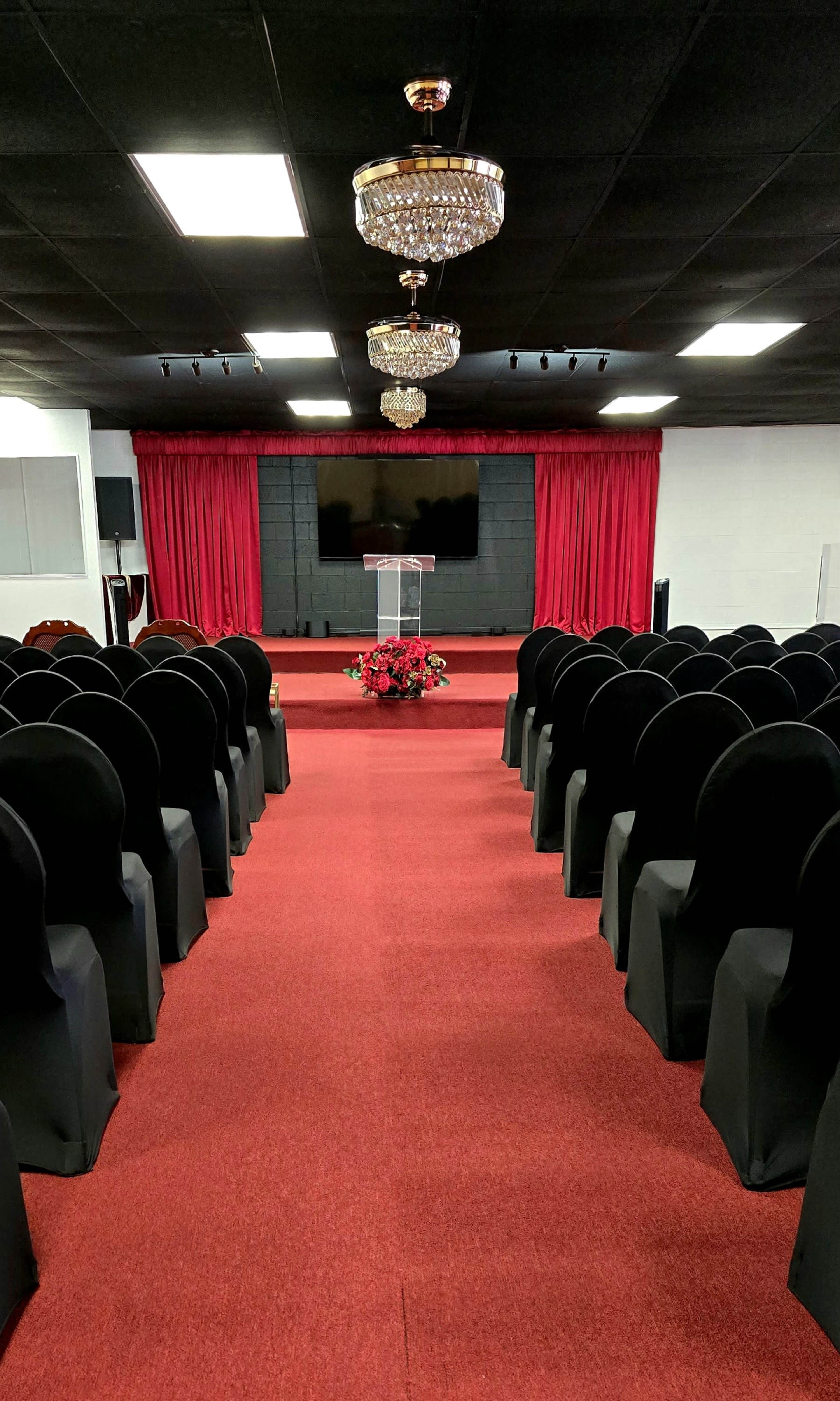 The image shows an empty auditorium with rows of black chairs, a red carpet, a decorative arrangement of flowers at the front, and a large screen above a podium.