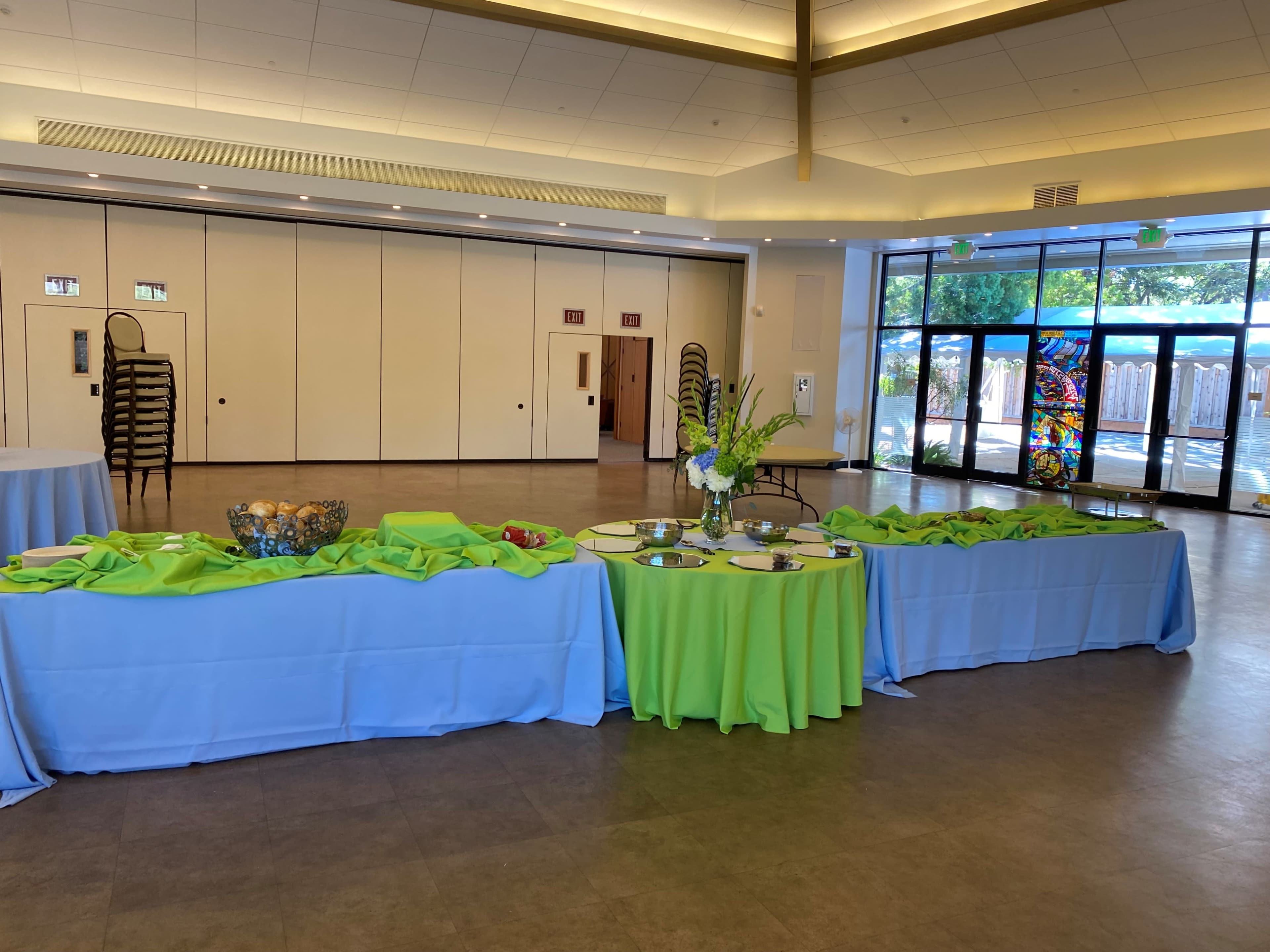 The image shows a spacious indoor venue set with tables covered in light blue and green tablecloths, arranged for an event, with decorations and a large window displaying natural light.