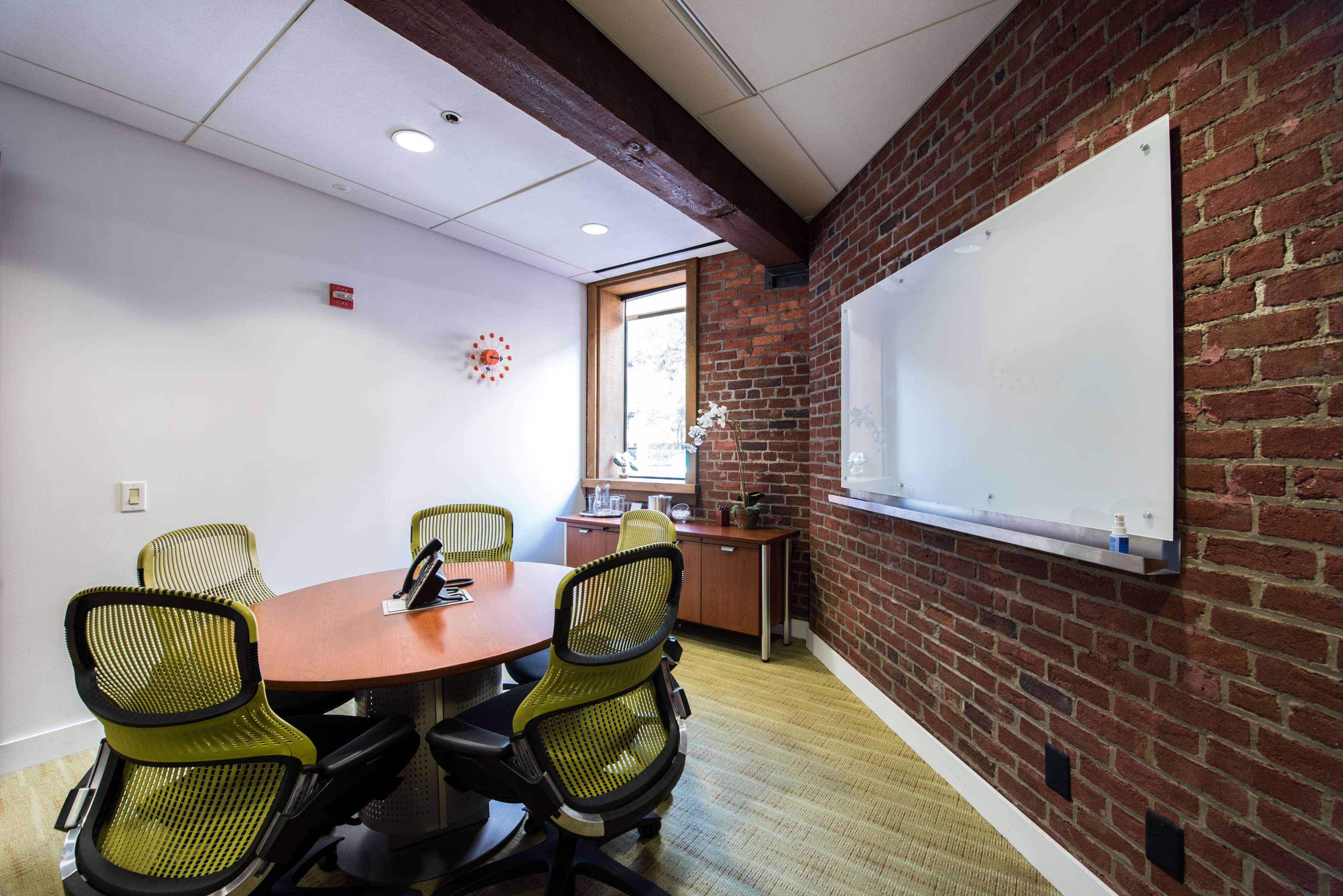 A conference room features a round table with four green chairs, a whiteboard on the wall, and a window with a view of the outside.