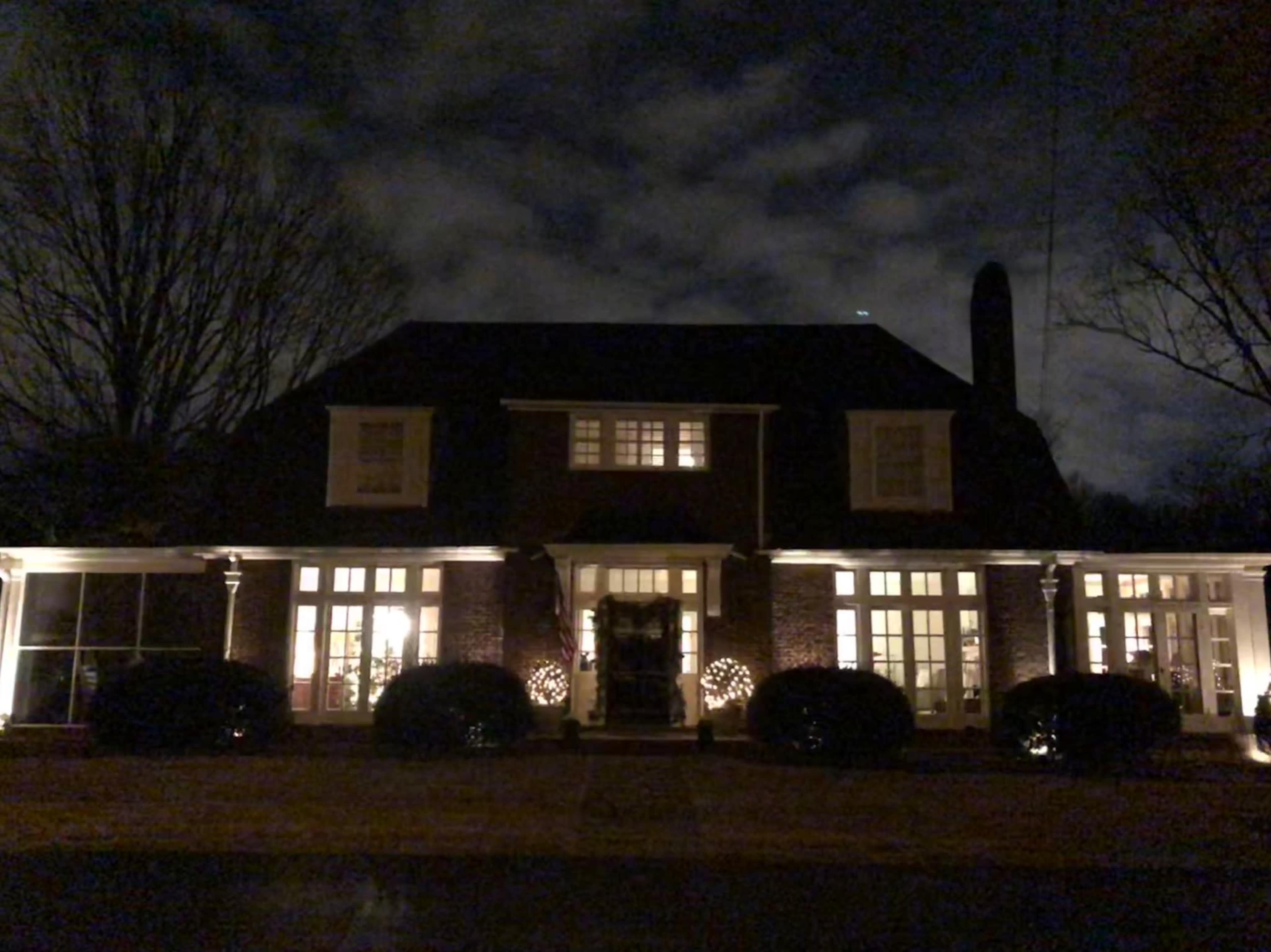 A two-story house is illuminated at night, featuring a symmetrical facade with large windows and decorative wreaths on either side of the front door.