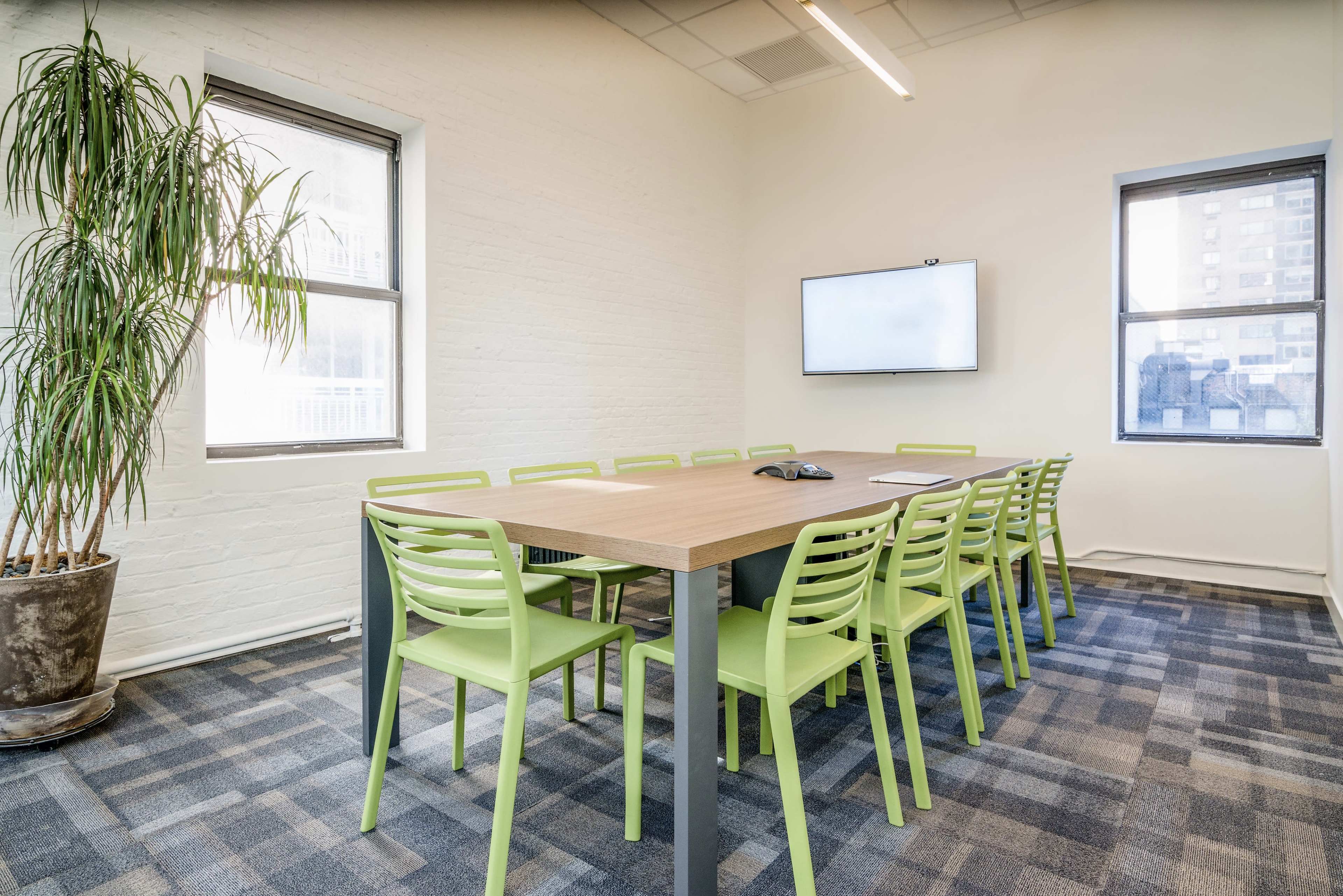 A meeting room features a long table surrounded by green chairs, with windows letting in natural light and a wall-mounted screen at one end.
