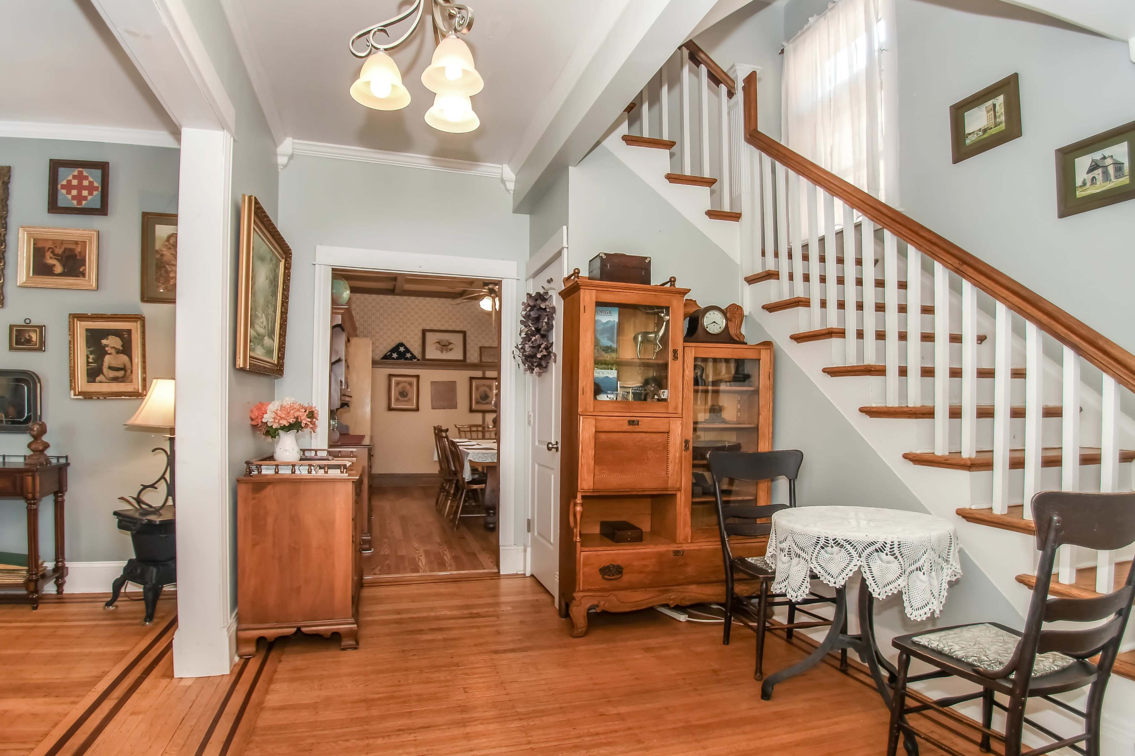 The image shows a well-lit entryway with a staircase, a small round table, and various framed pictures on the walls.
