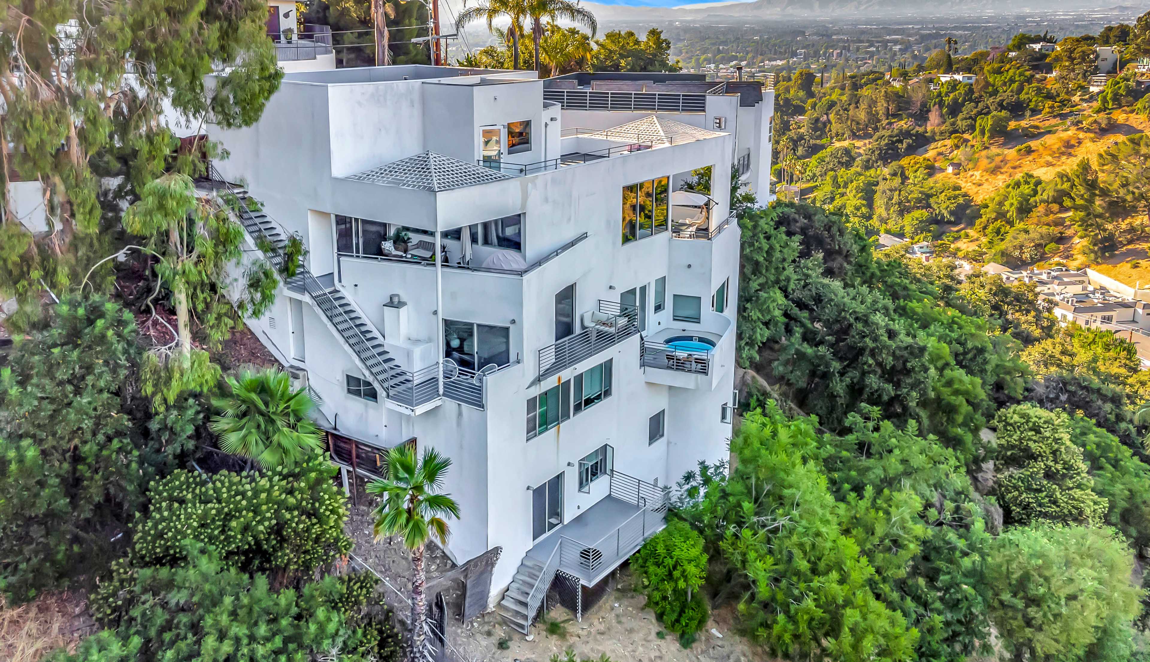 A modern, multi-level white house is situated on a hillside, surrounded by trees, with outdoor staircases and a rooftop area.