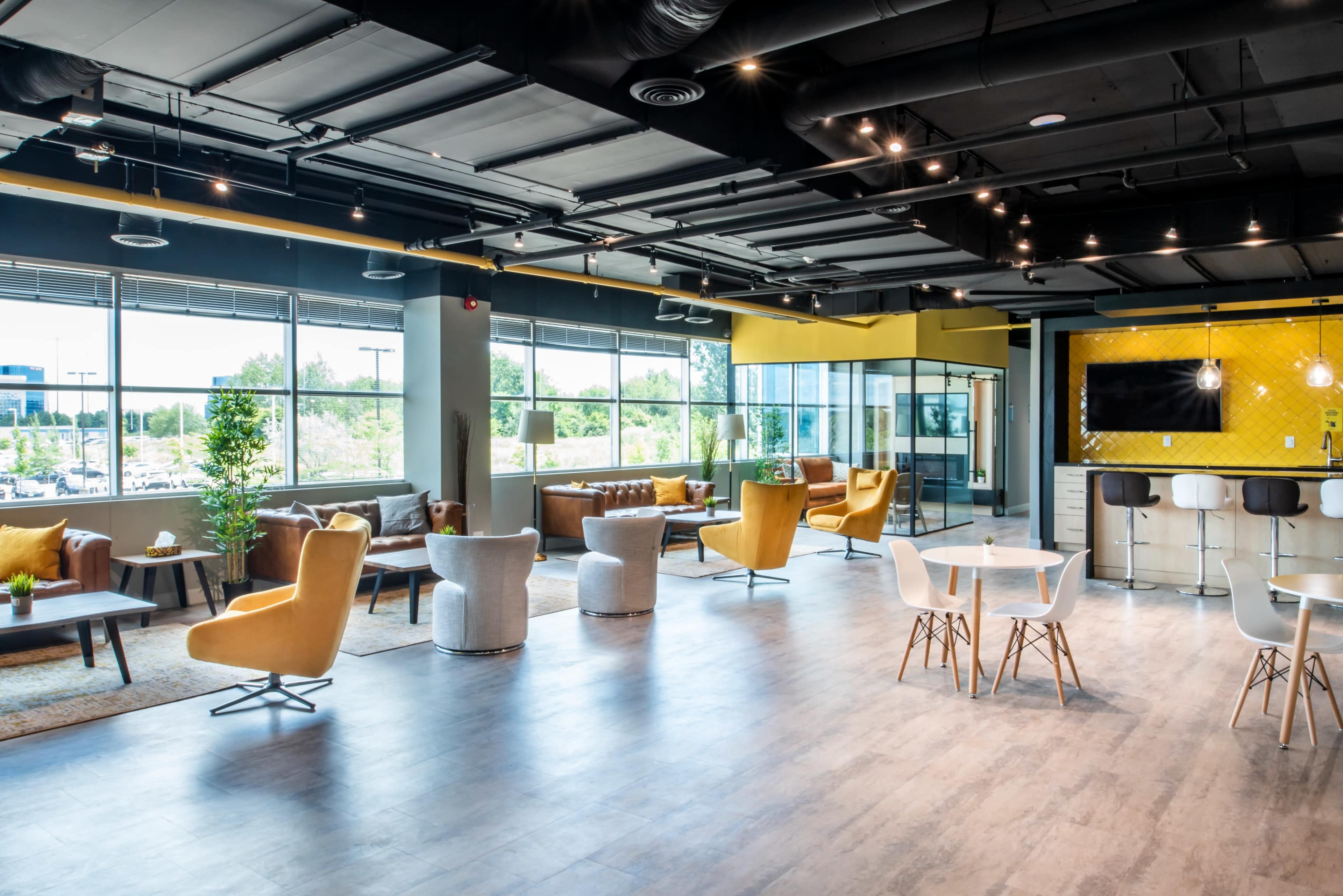 A modern office lounge area with various seating options, large windows, and a kitchen space in the background.