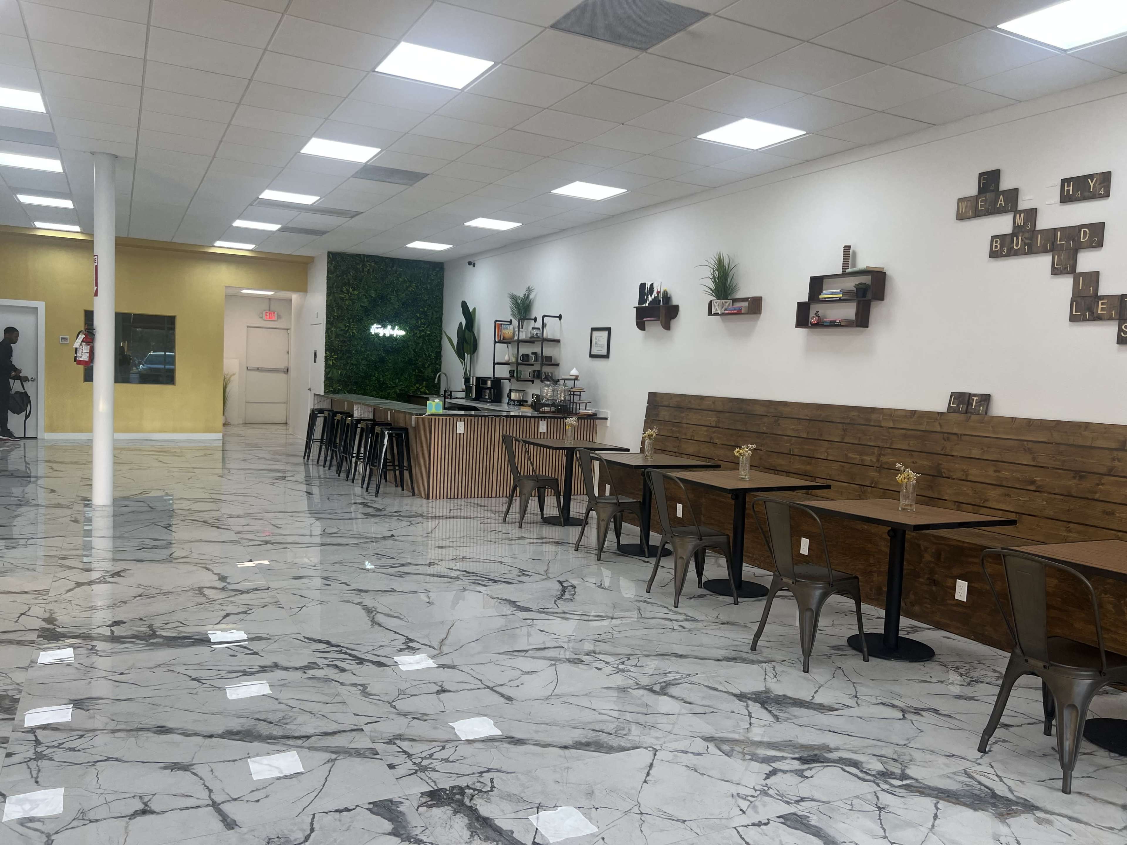 The image shows a modern café interior with marble floors, wooden seating arrangements, a counter displaying refreshments, and greenery on the walls.