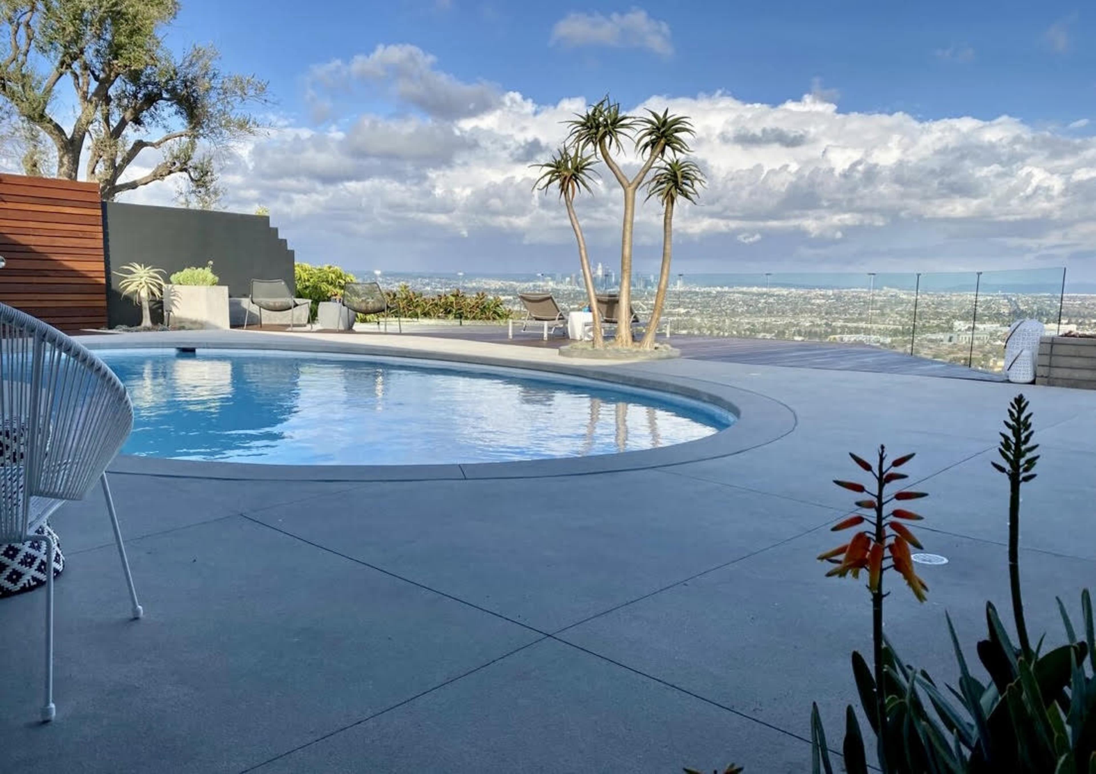 A swimming pool with a curved edge, surrounded by a concrete patio and overlooking a city skyline beneath a cloudy sky.