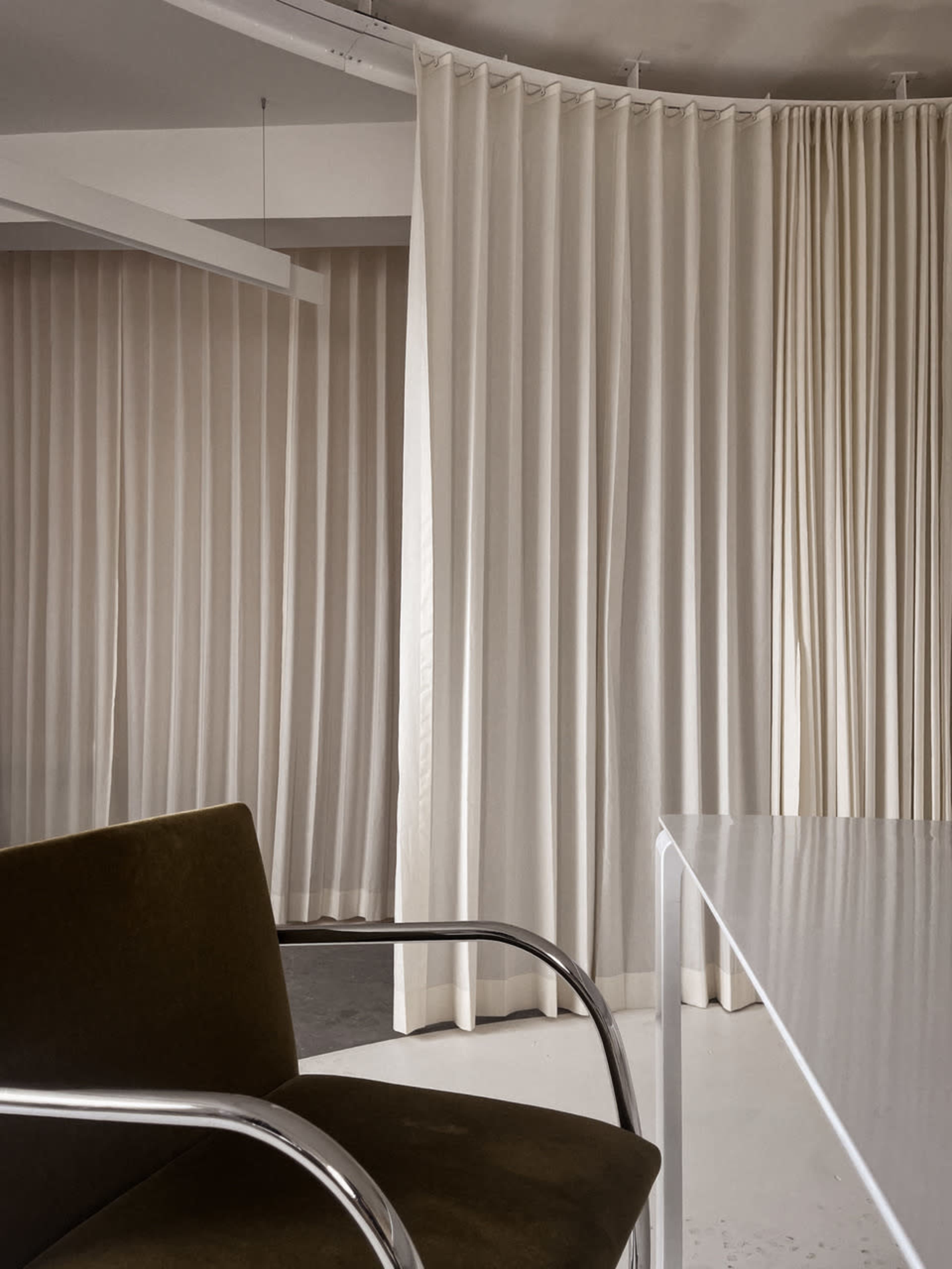 The image shows a modern interior space featuring light-colored draped curtains and a minimalist chair with a chrome frame.