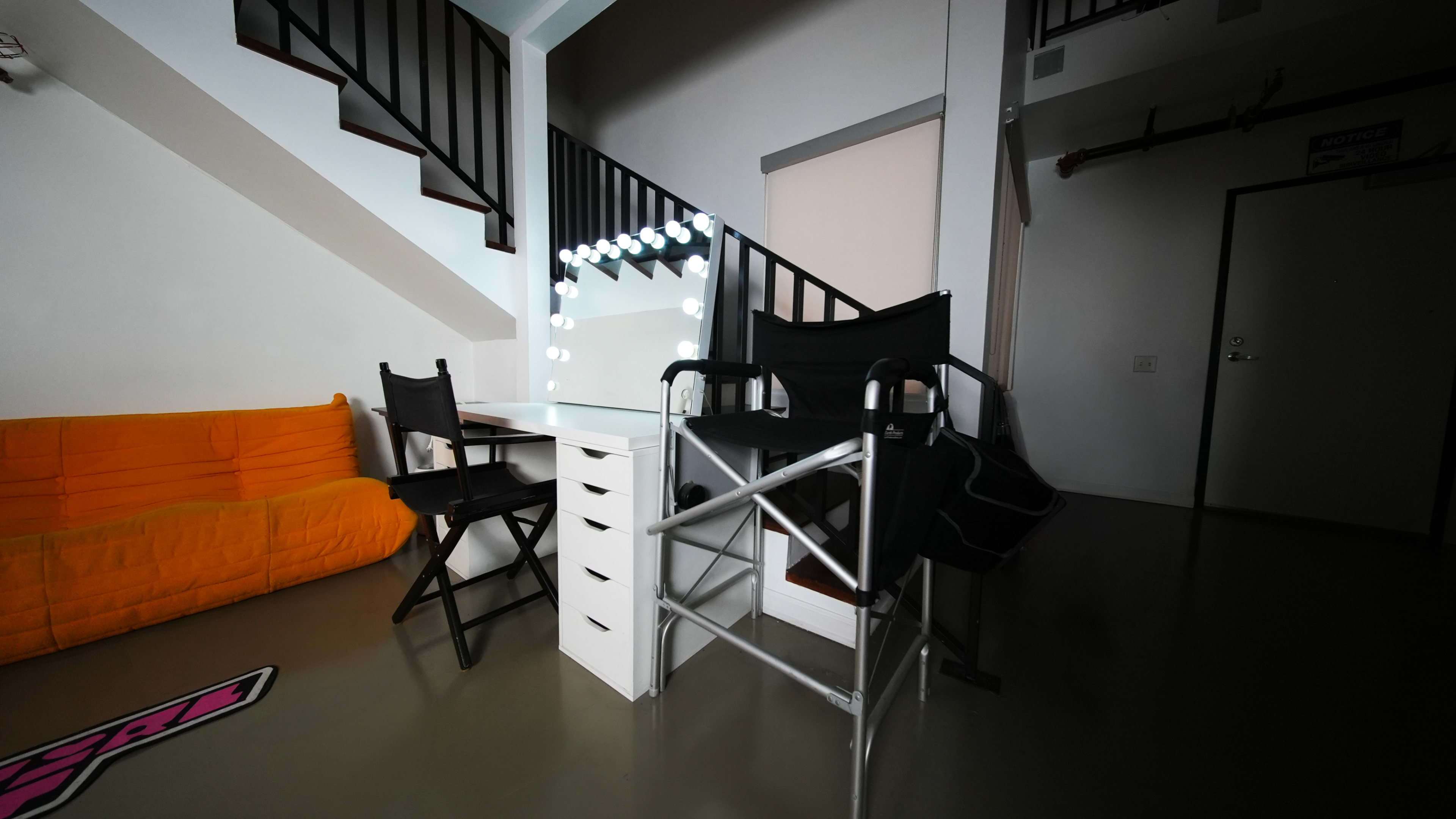 The image shows a modern room featuring a bright orange sofa, a vanity table with lights, and two tall director's chairs.