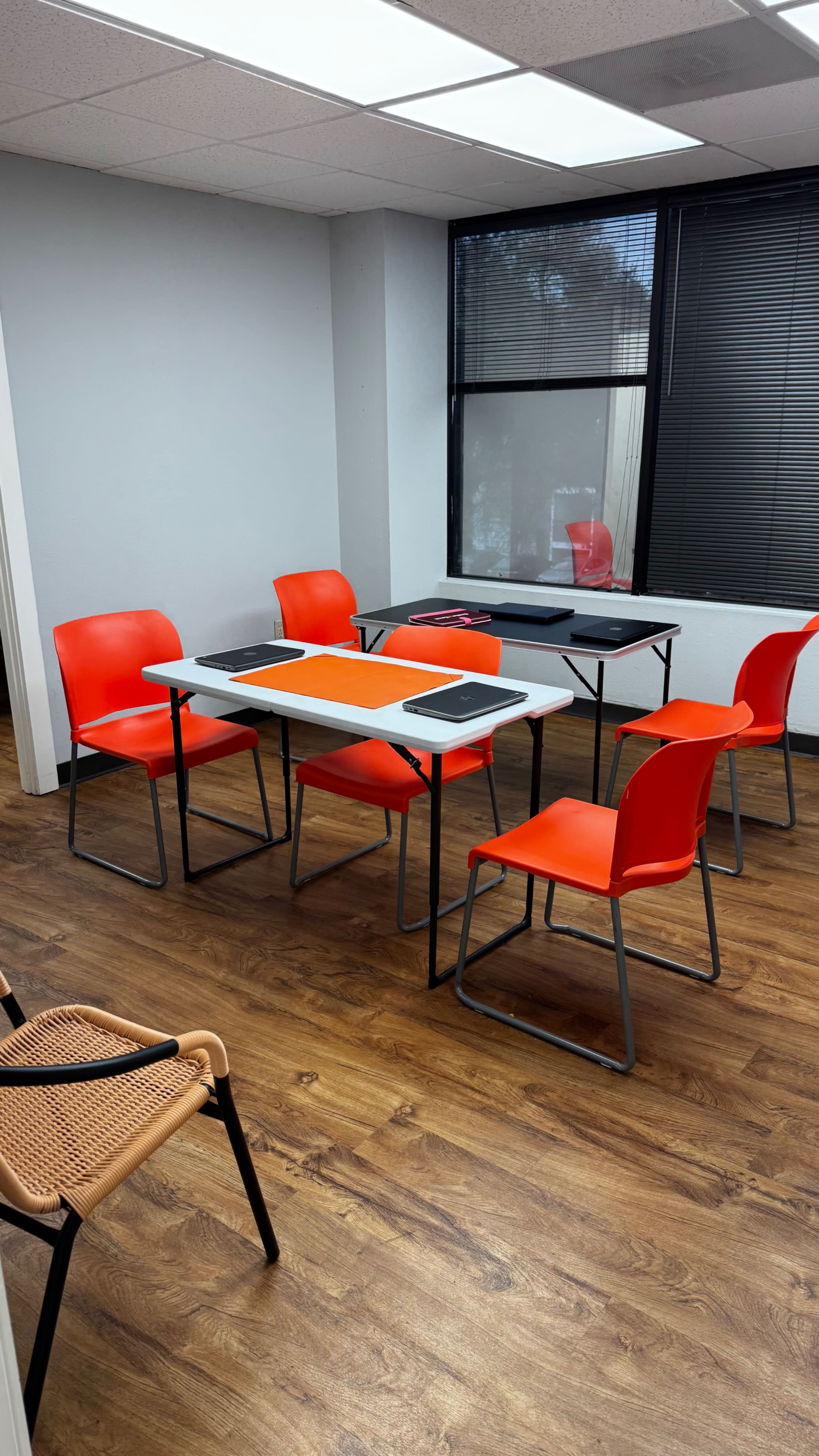 The image shows a small meeting room with a rectangular table surrounded by six orange chairs and a large window.