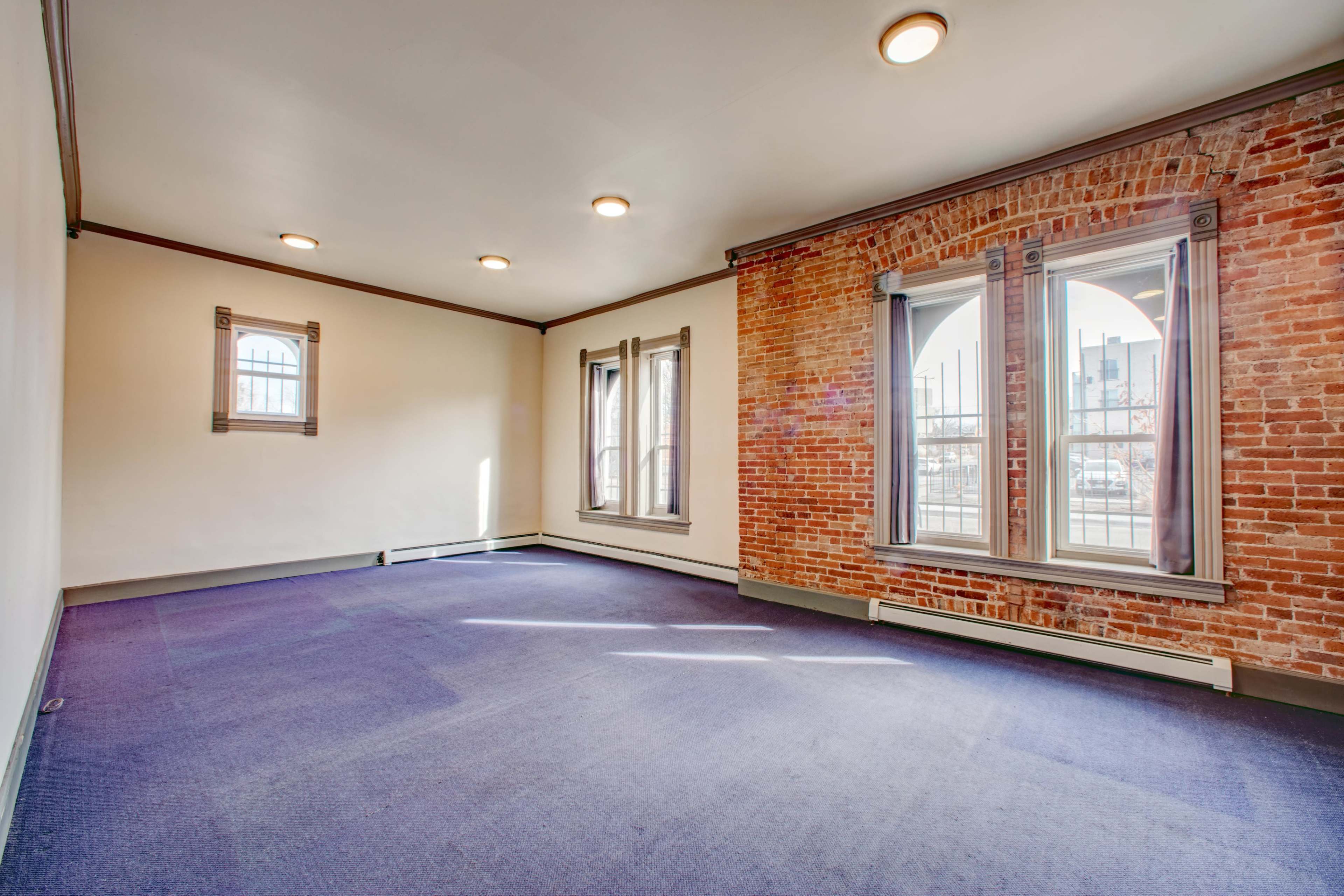 The room features exposed brick walls, two large windows, and a carpeted floor with overhead lighting.