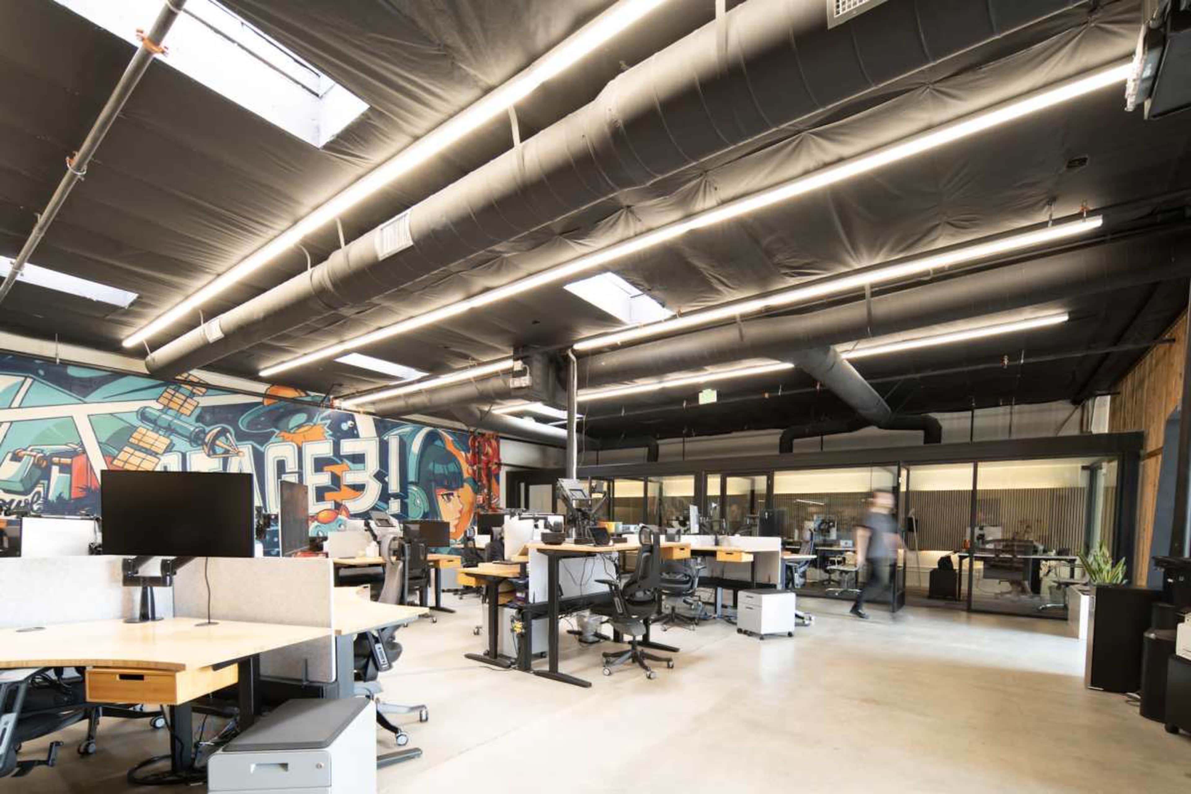 The image shows a modern office space with several workstations and a large mural on one wall.