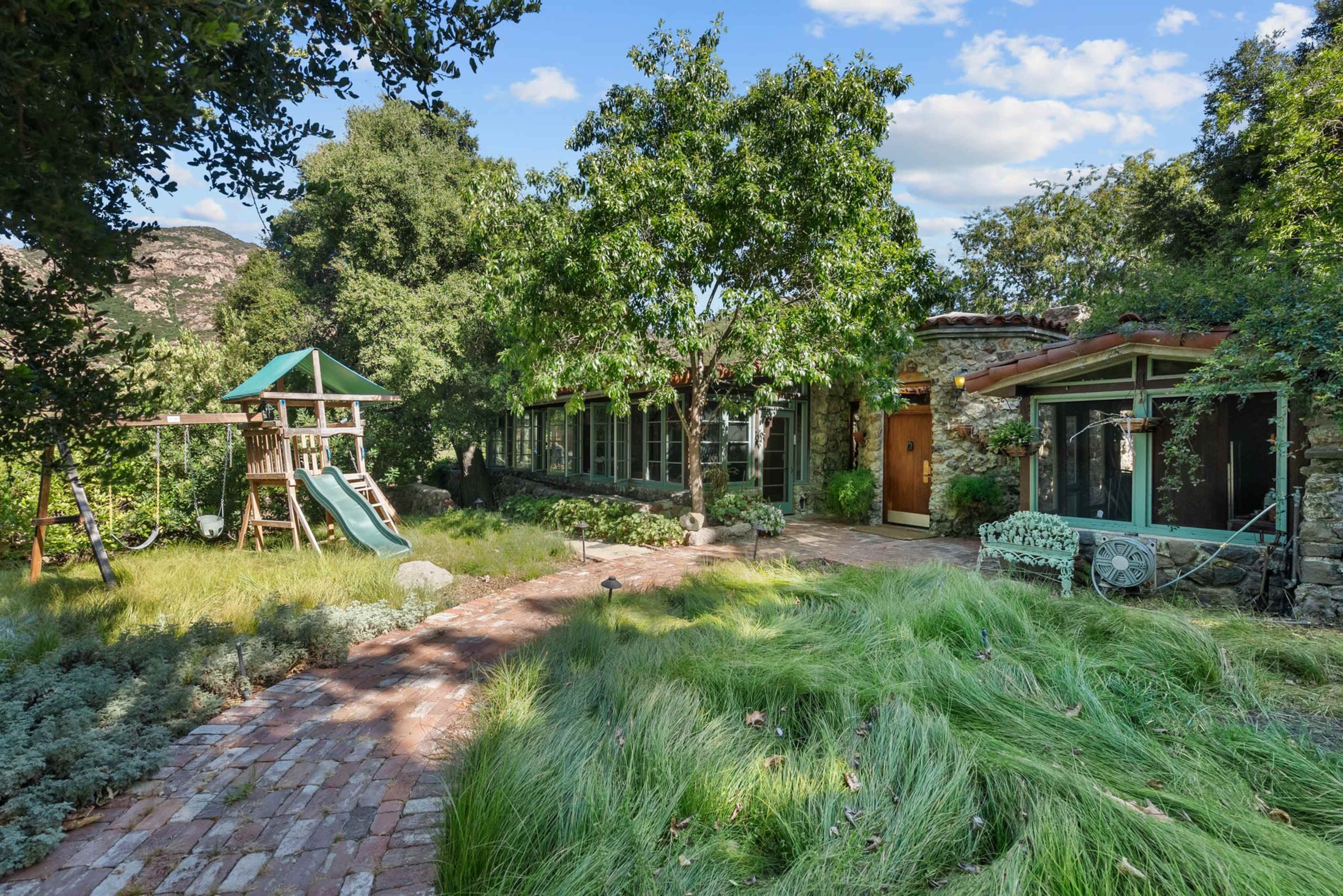 The image shows a scenic backyard featuring a stone house, a children's playset with a slide, and landscaped greenery.