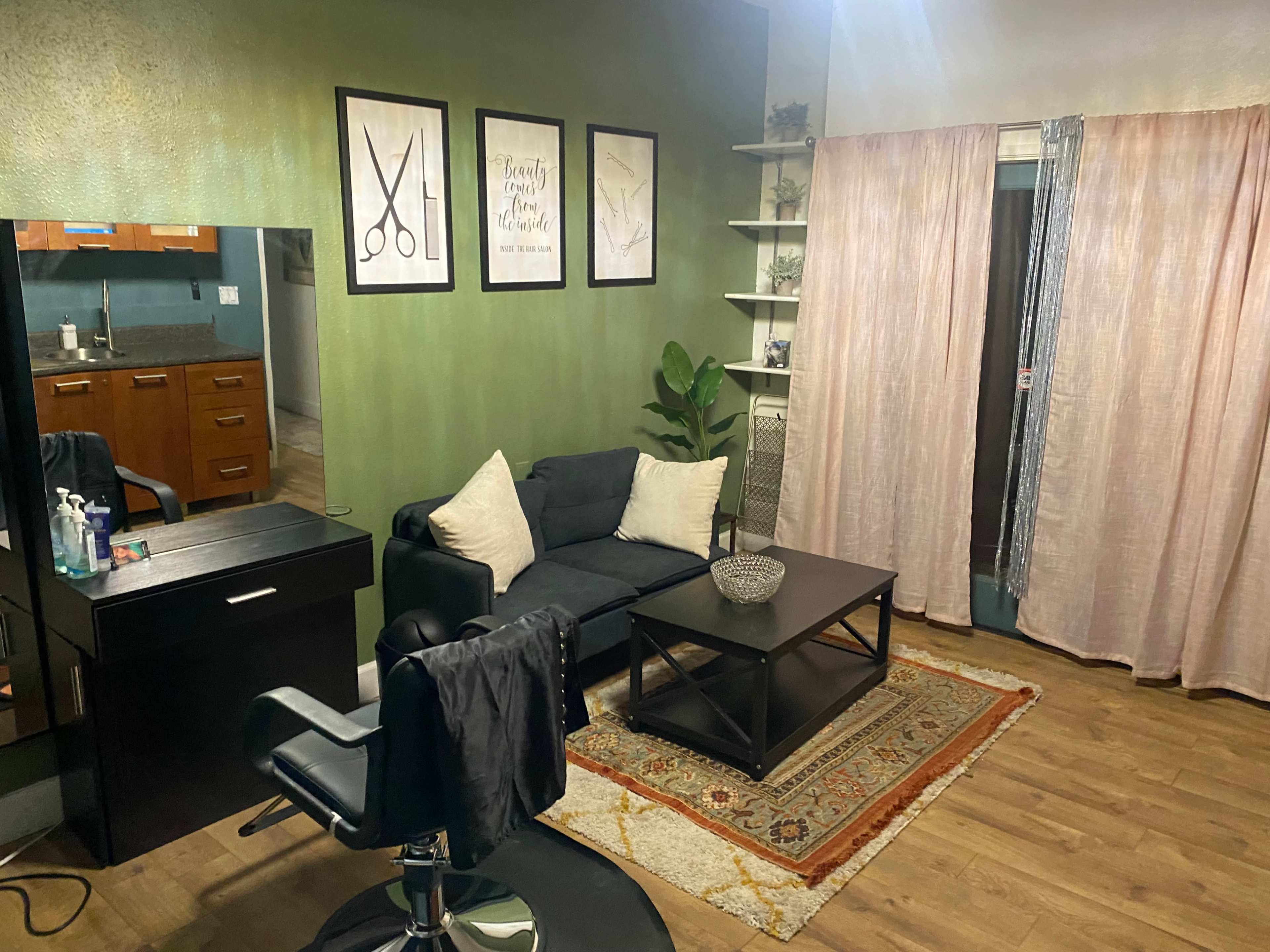 A small hair salon interior with a black sofa, a coffee table, a stylist's chair, and green walls adorned with framed scissors art.