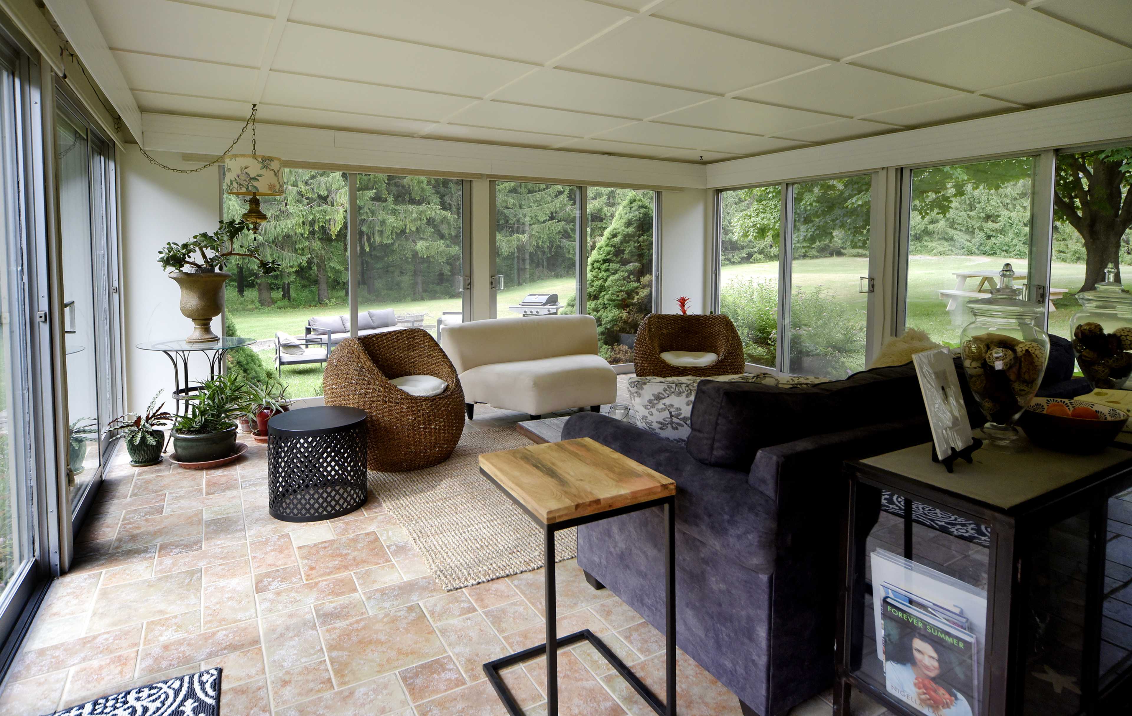 A sunlit indoor seating area with large windows, featuring a mix of wicker and fabric furniture, potted plants, and a view of a grassy outdoor space.