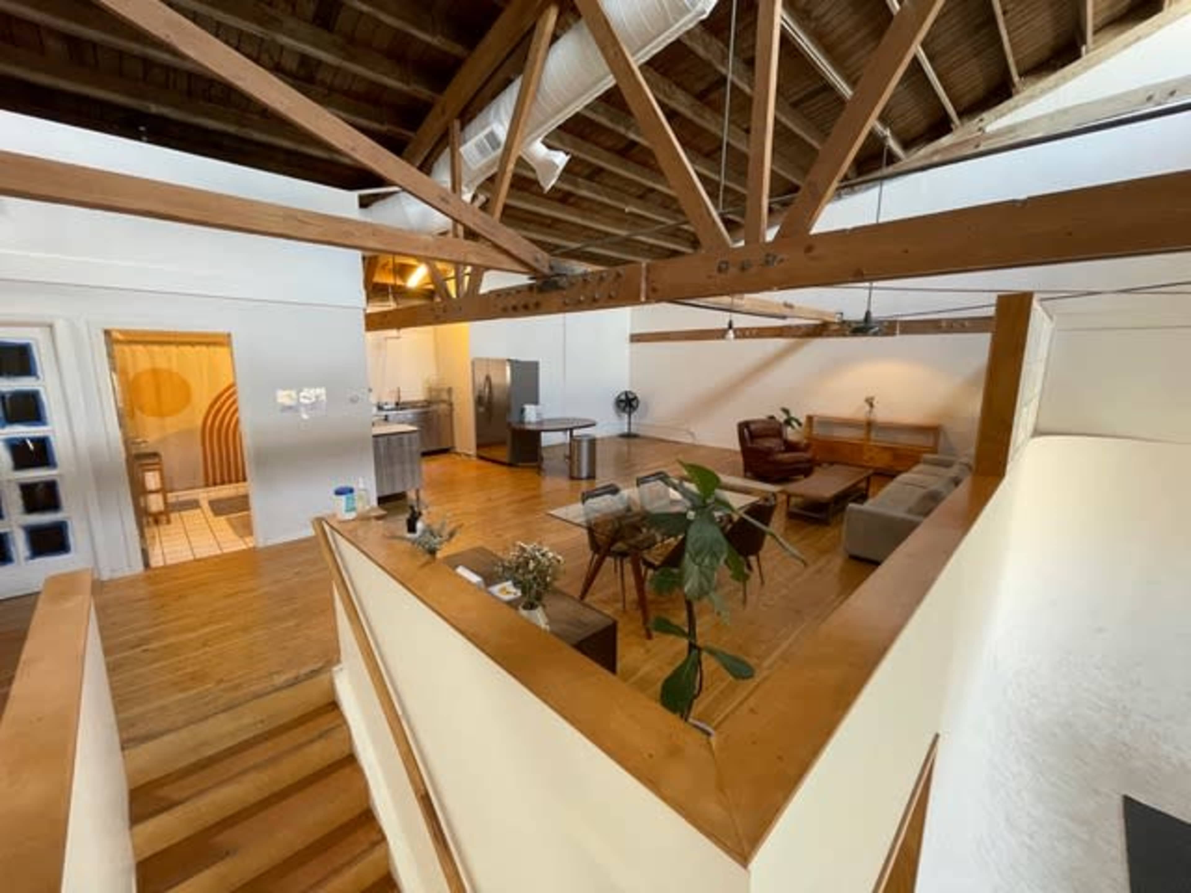 The image shows a spacious interior with wooden beams, featuring a living area with a sofa, a dining table, and a kitchen in the background.
