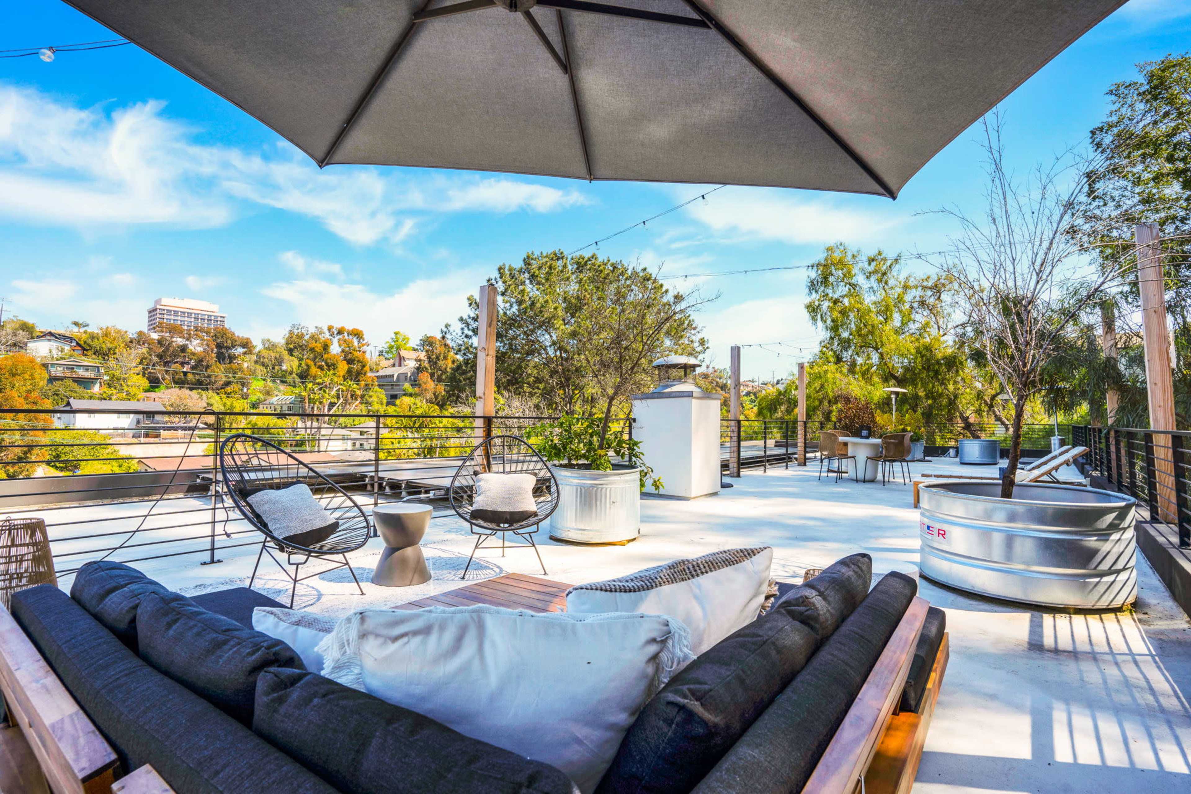 A rooftop patio featuring a sectional sofa, two chairs, an umbrella, and a view of surrounding greenery.