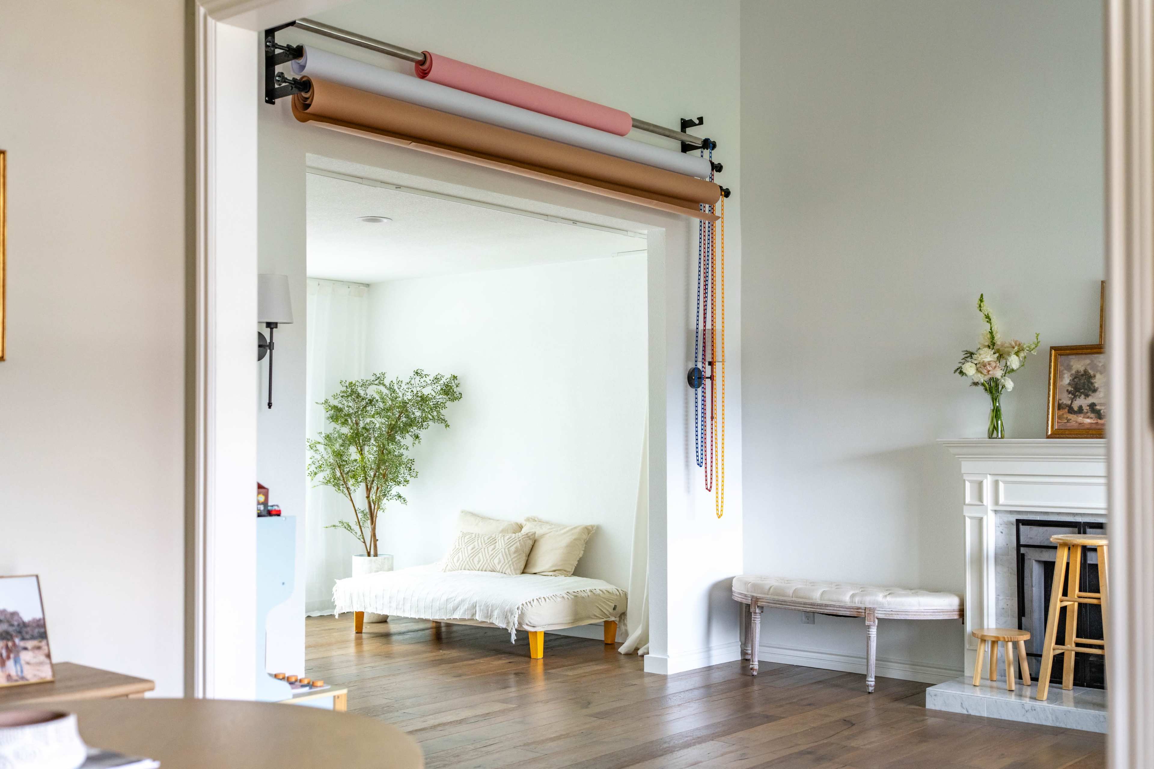 The image shows a well-lit room divided by an open doorway, featuring a comfortable daybed and a wall-mounted system for hanging rolls of paper in various colors.