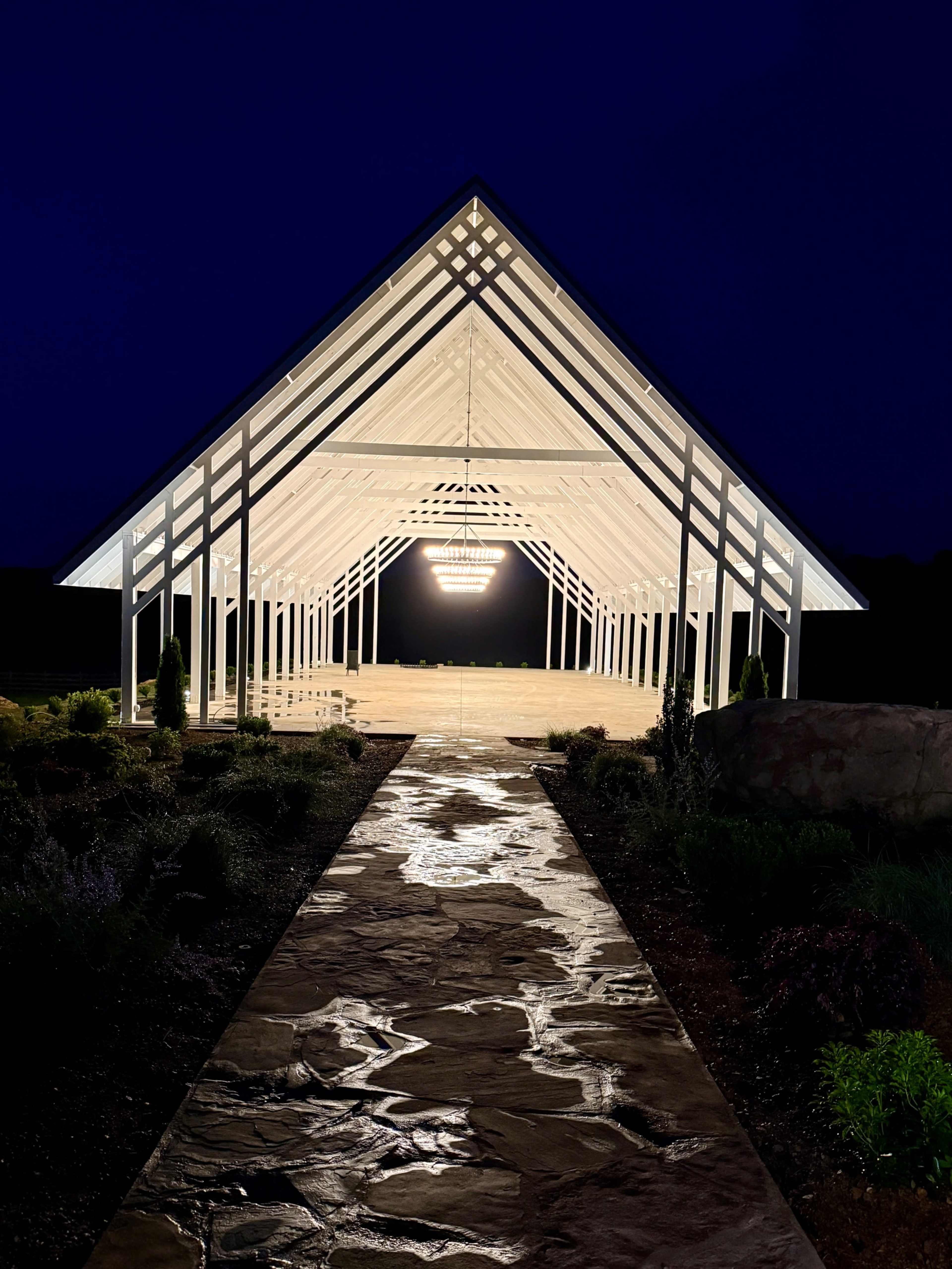 A modern, illuminated barn-style structure stands at night, flanked by a stone path and surrounded by landscaped greenery.