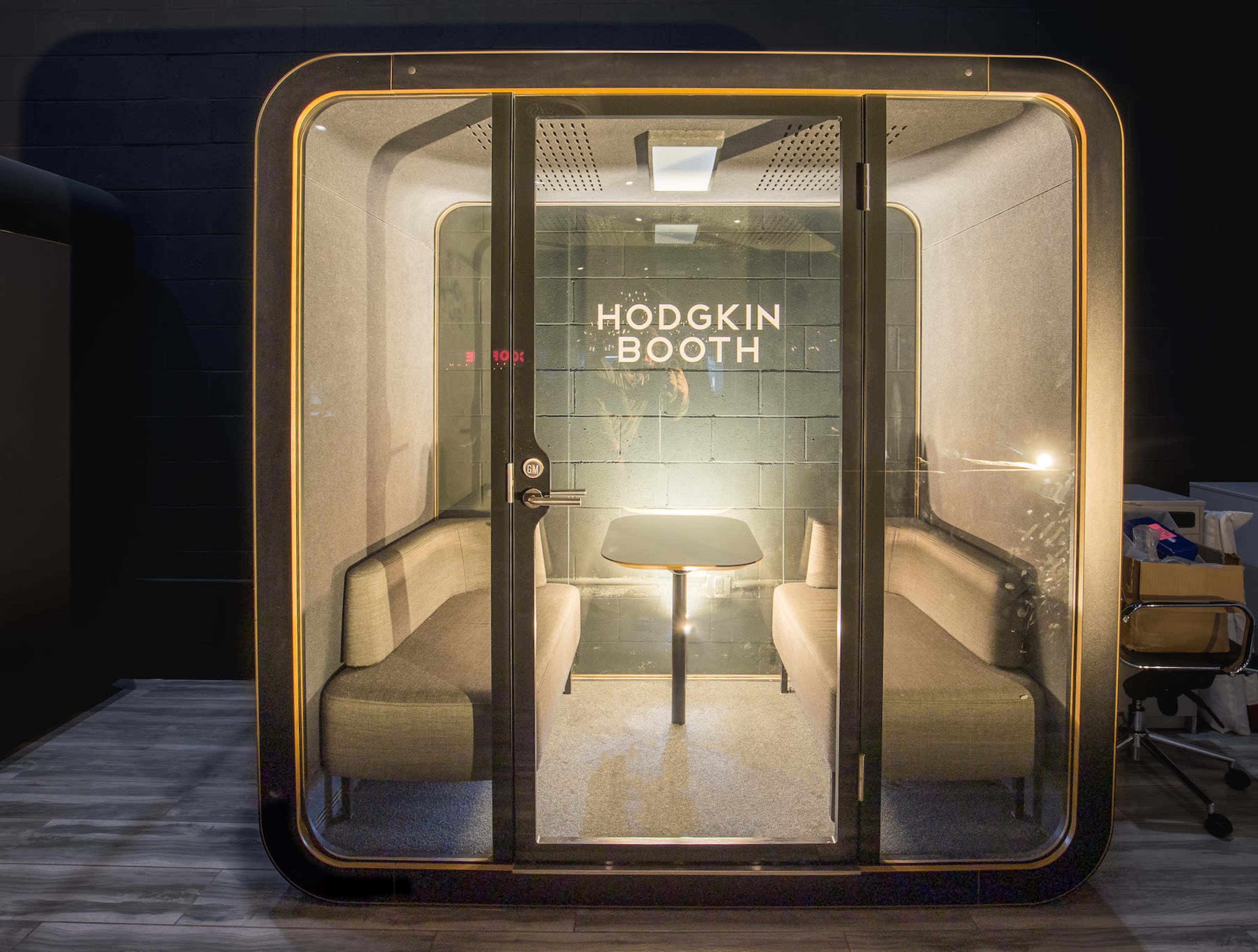 The image shows a modern soundproof booth with a glass exterior, featuring two benches and a small table inside, labeled "HODGKIN BOOTH."