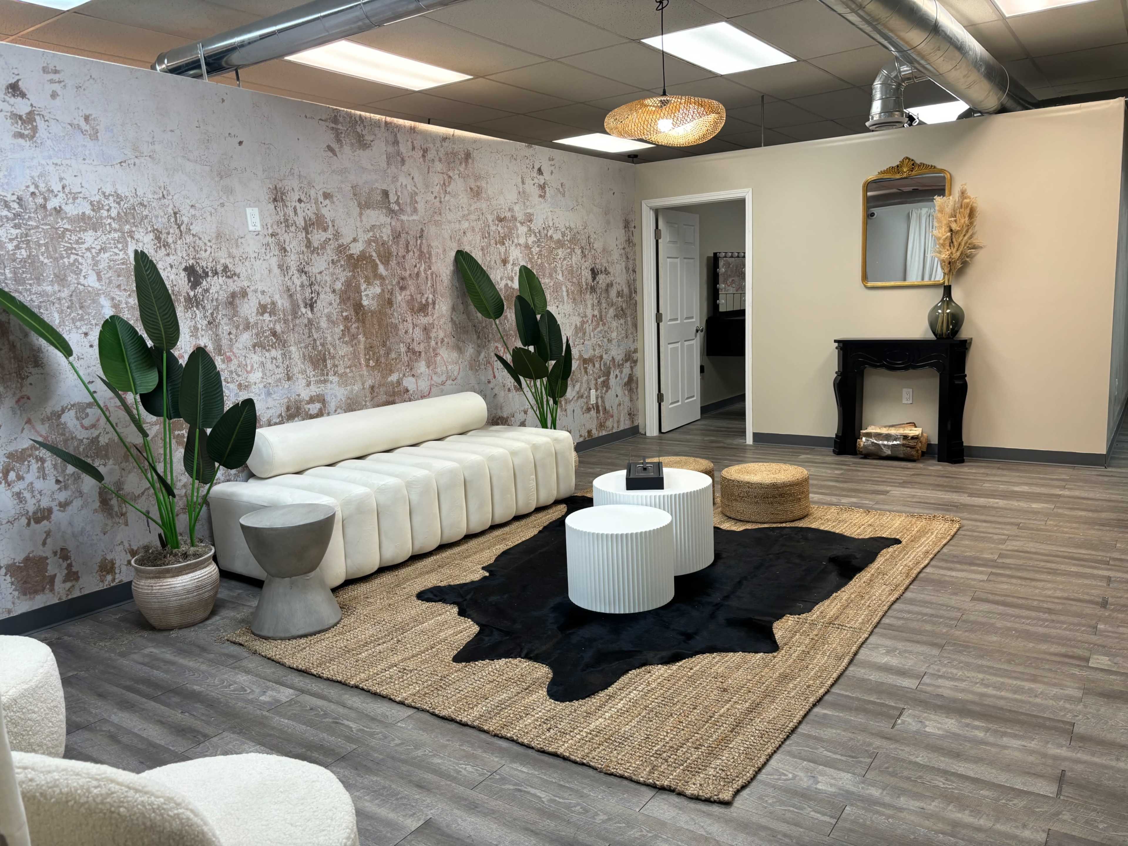 The image shows a modern lounge area featuring a white sofa, two small round tables, a woven rug, and decorative plants against a textured wall.
