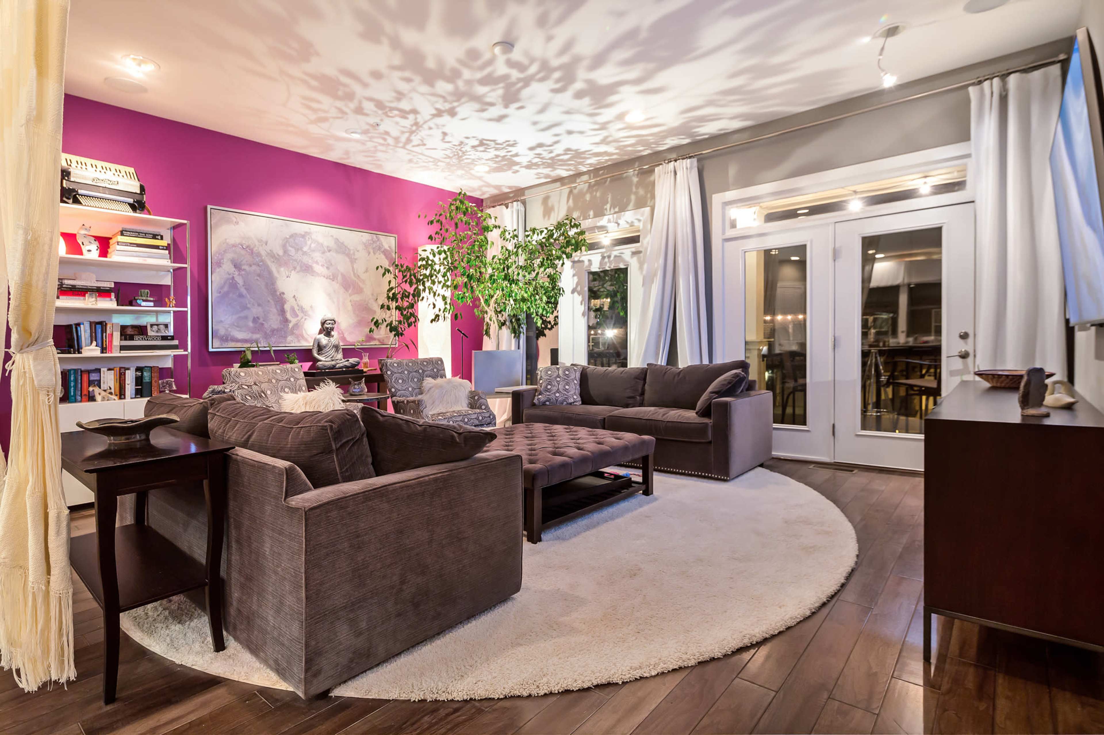 The living room features a circular rug surrounded by gray sofas, a coffee table, and a large potted plant, with a purple accent wall and glass doors leading outside.