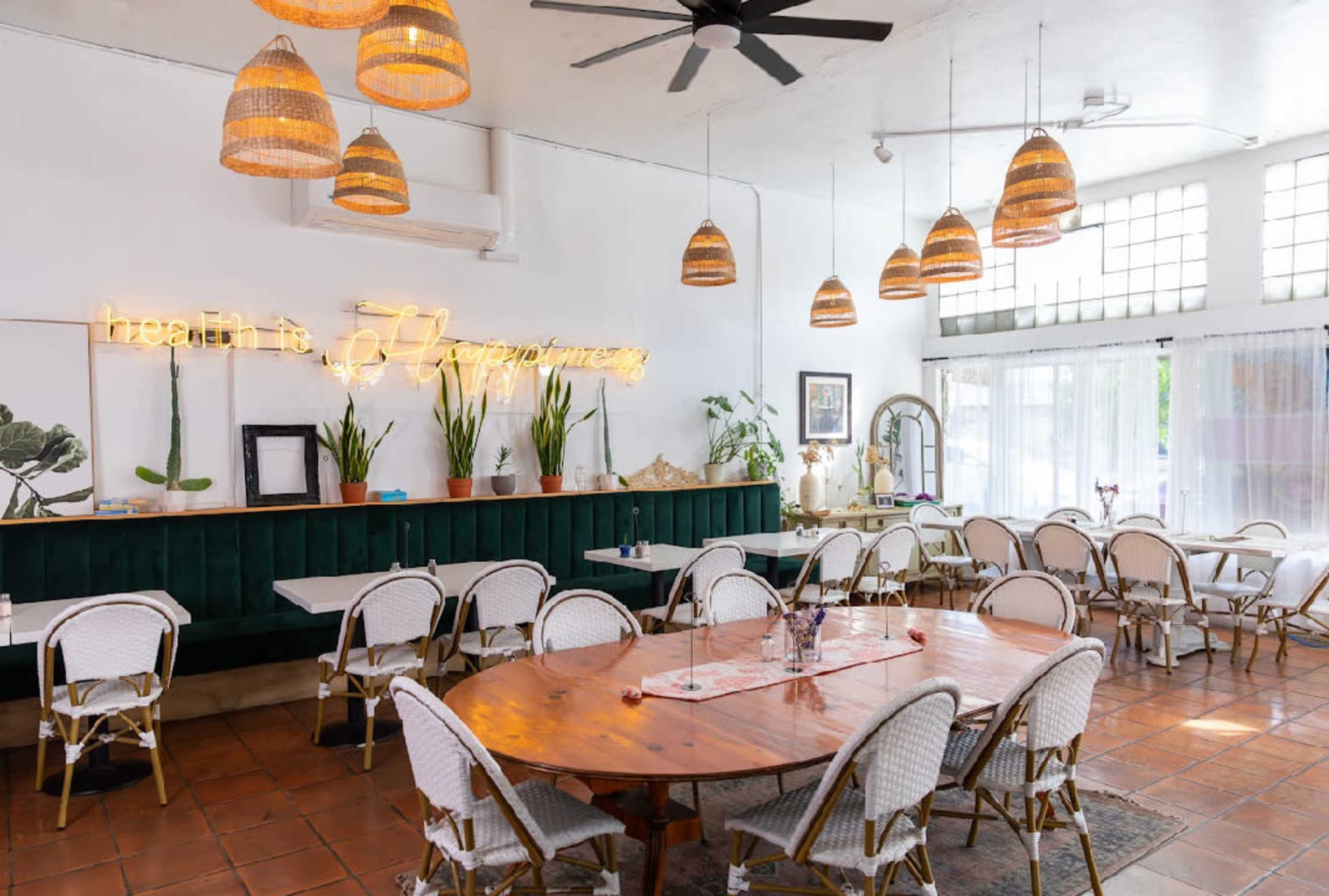 The image shows a bright and spacious dining area with wooden tables, wicker chairs, and hanging pendant lights, featuring green accents and large windows.