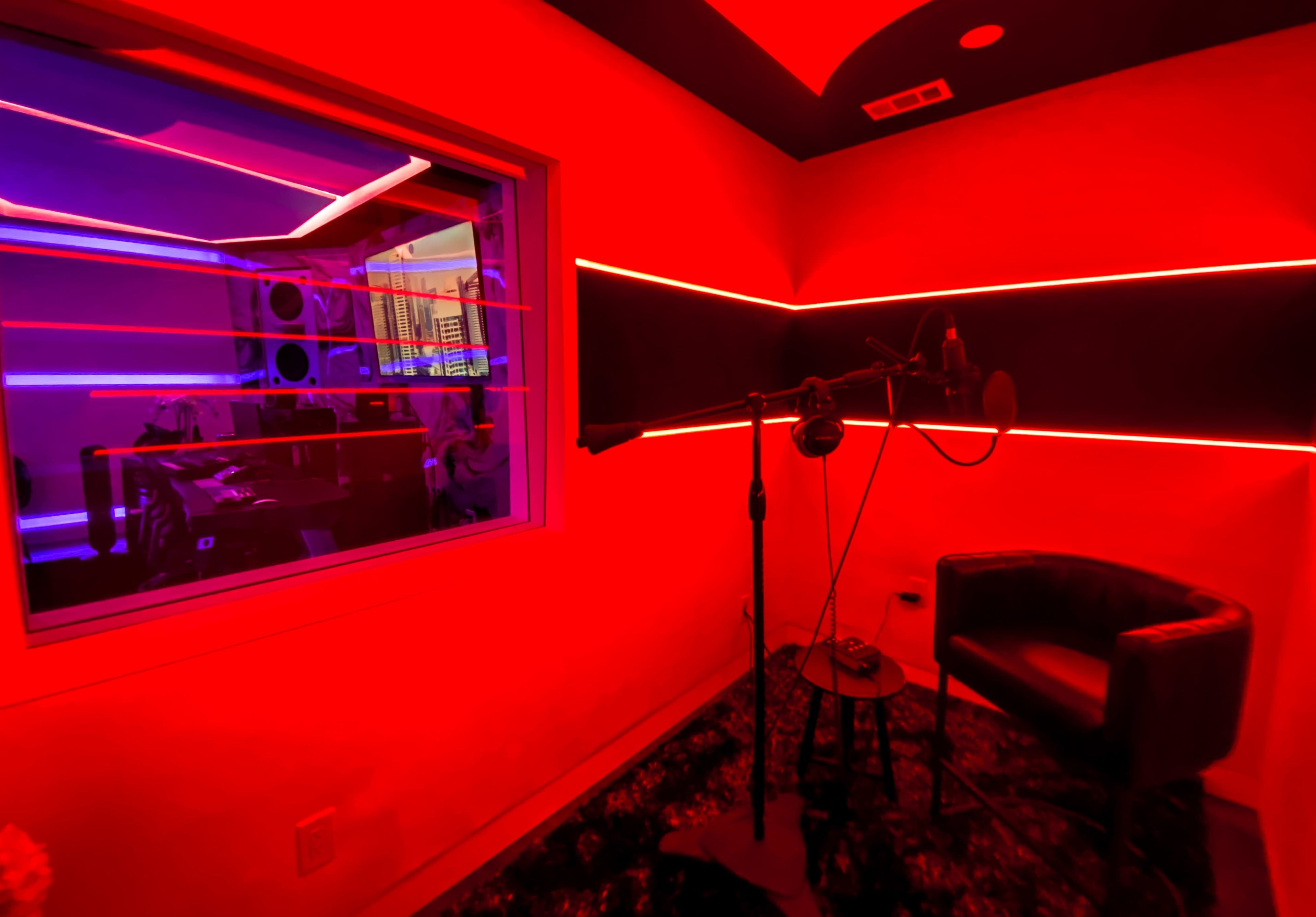A recording studio features a microphone stand and a chair in a room illuminated by red lighting, with a window revealing equipment in an adjacent space.