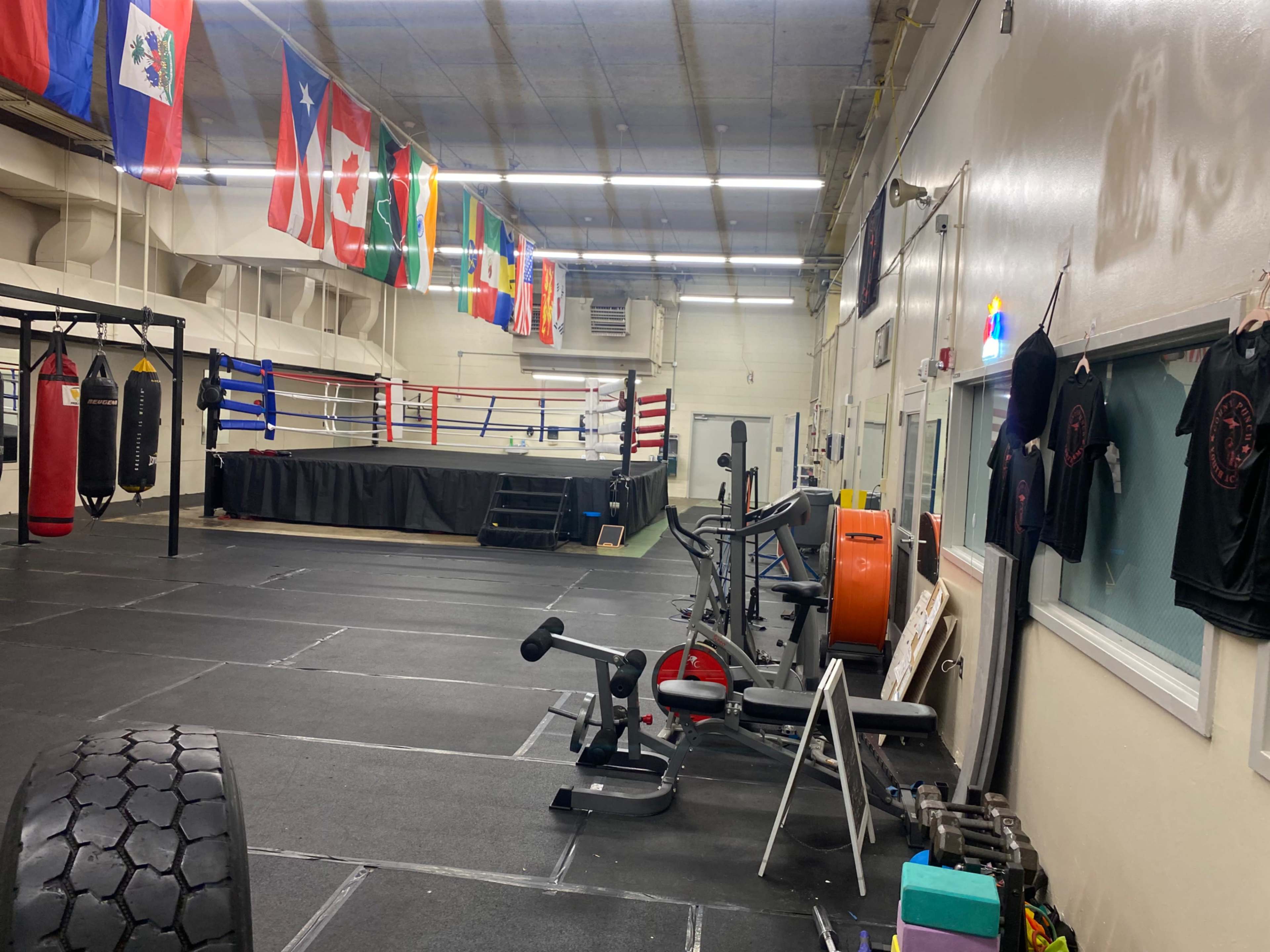 The image shows a boxing gym with various exercise equipment, a boxing ring in the background, and several flags hanging from the ceiling.