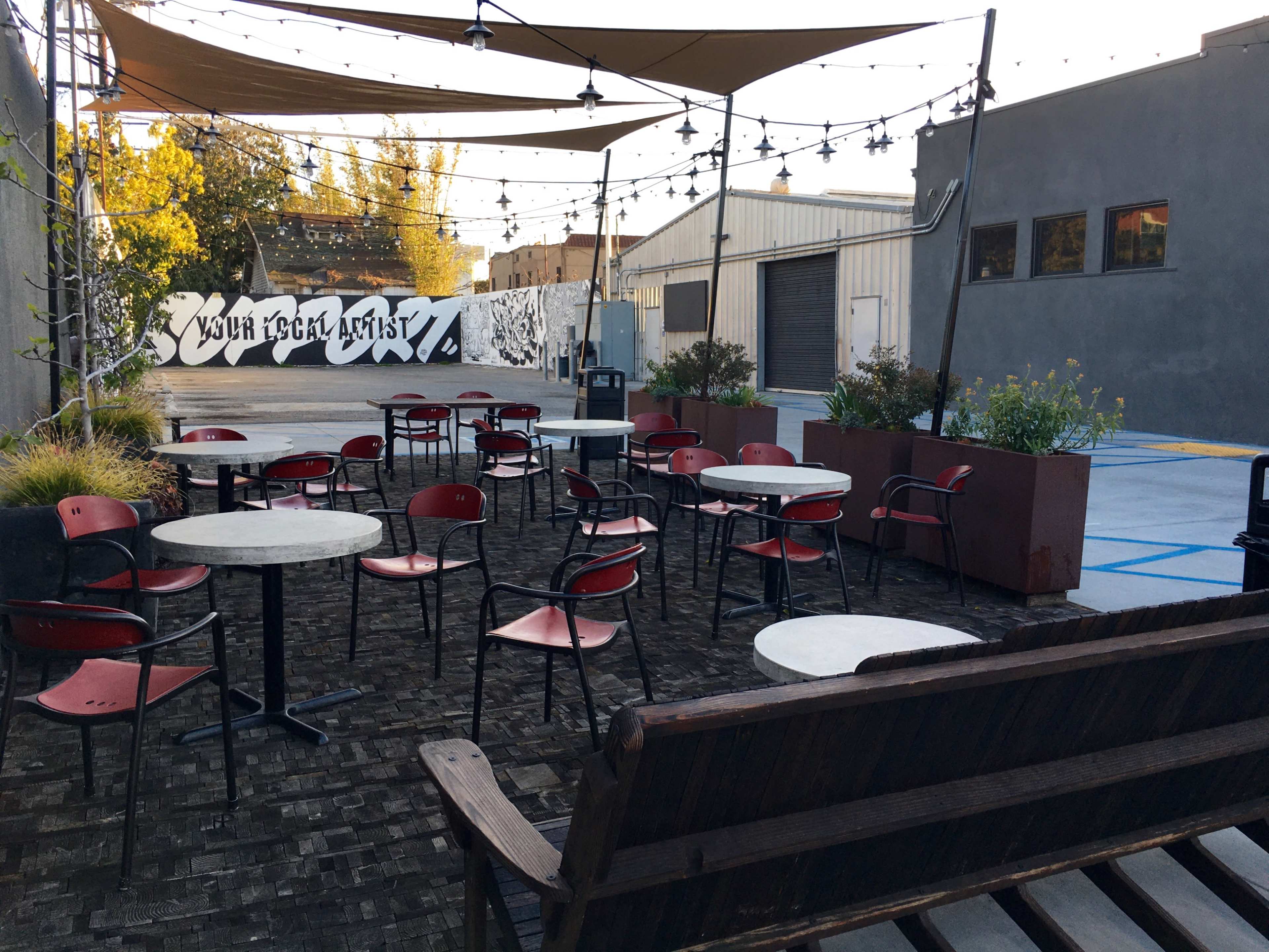 An outdoor seating area features red metal chairs and small tables under a canopy with string lights, adjacent to a mural wall and a parking lot.