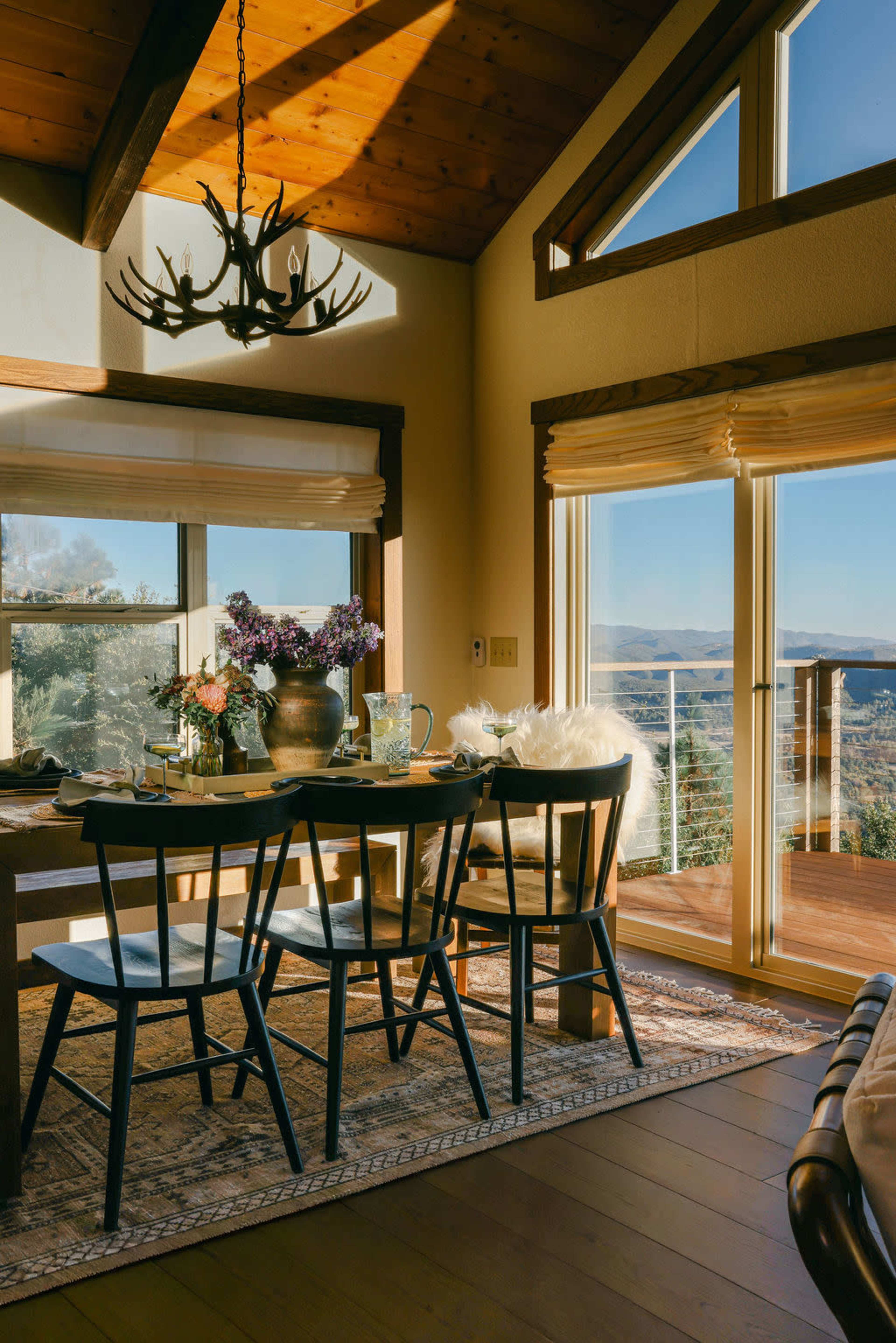 A dining area features a wooden table surrounded by black chairs, illuminated by sunlight streaming through large windows with scenic views.