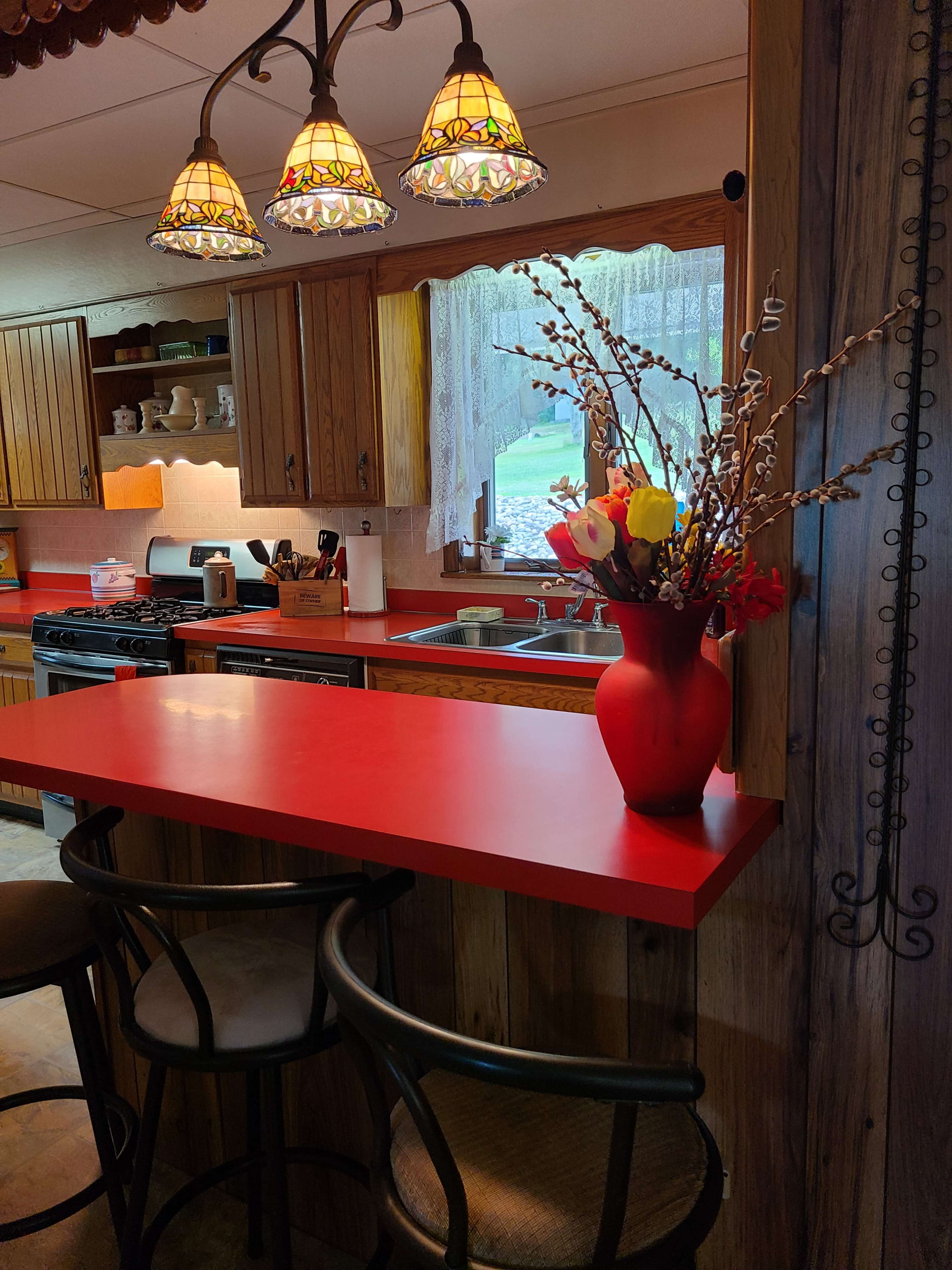 A kitchen with wooden cabinets, a red countertop, and a vase of flowers on the bar, illuminated by stained glass pendant lights.