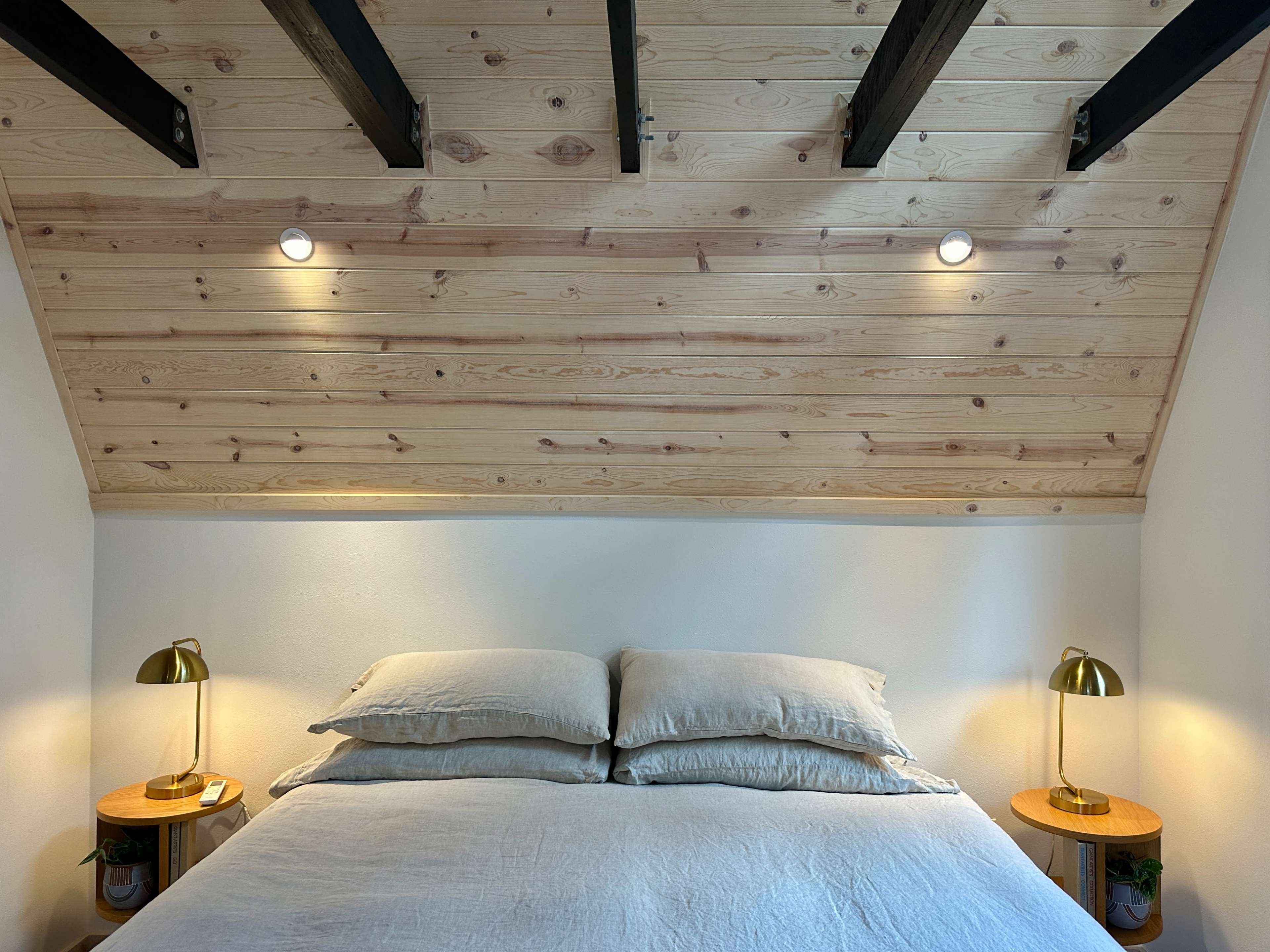 A neatly made bed with light gray pillows and blankets is positioned against a wooden slatted wall, flanked by two bedside lamps.