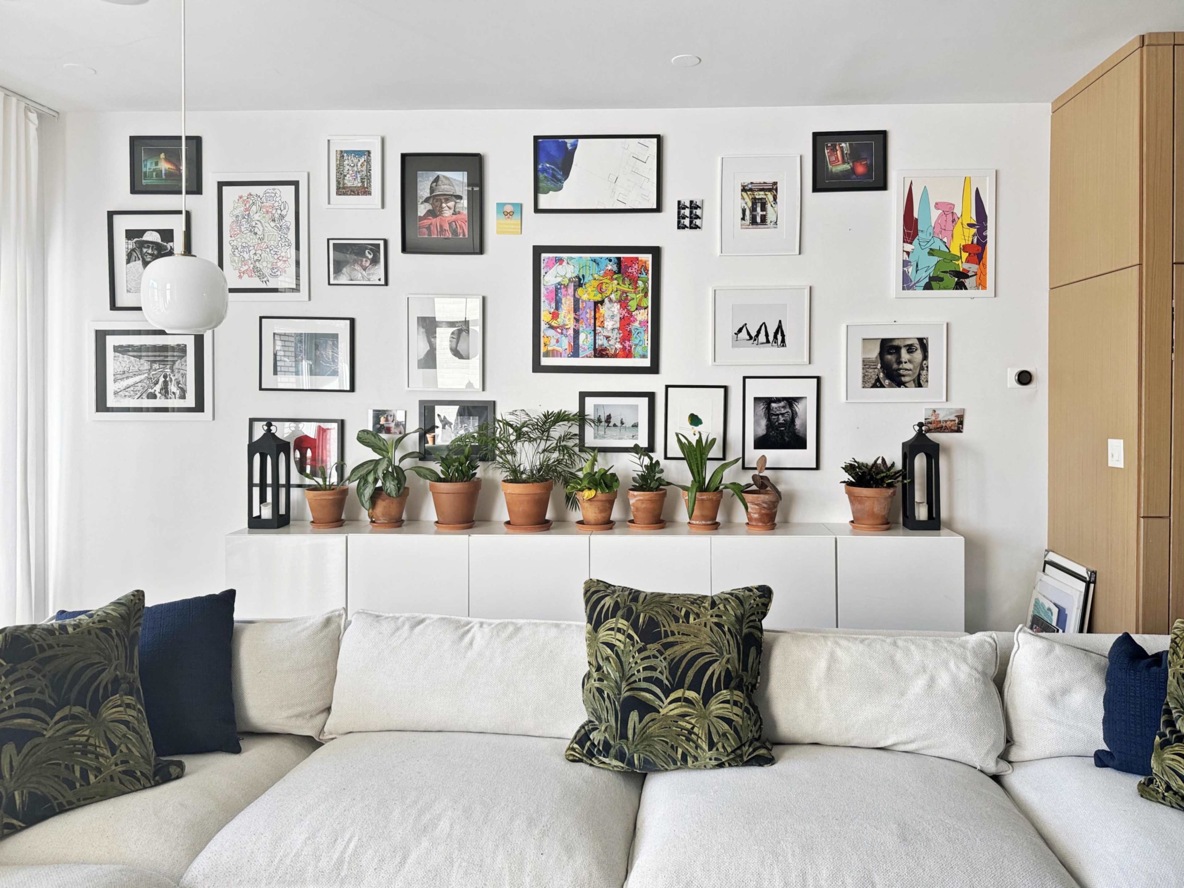 A living room wall adorned with various framed artworks surrounded by potted plants on a white shelf, with a light-colored, upholstered sectional sofa in the foreground.