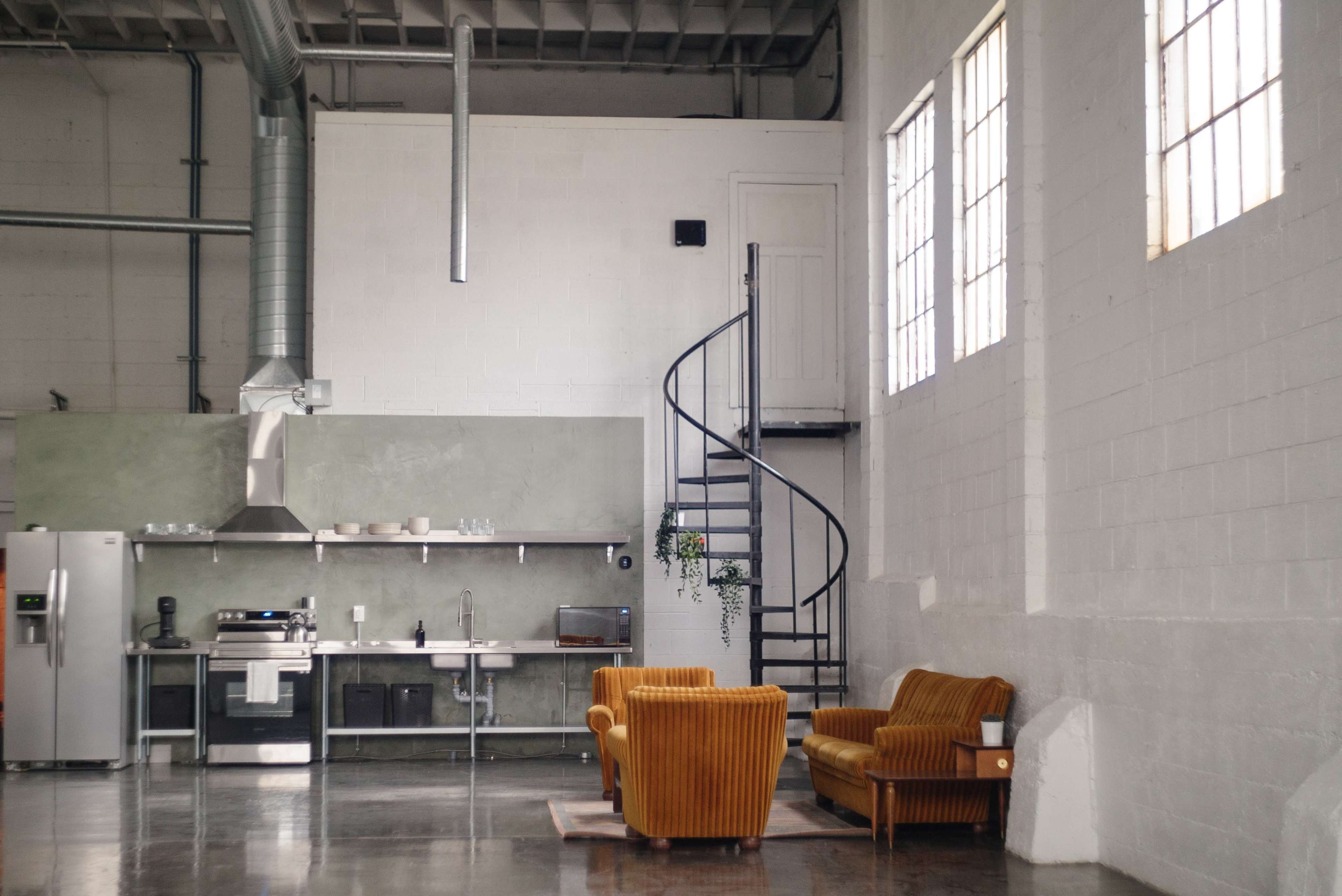A modern kitchen with a spiral staircase, featuring two orange chairs, large windows, and industrial elements.