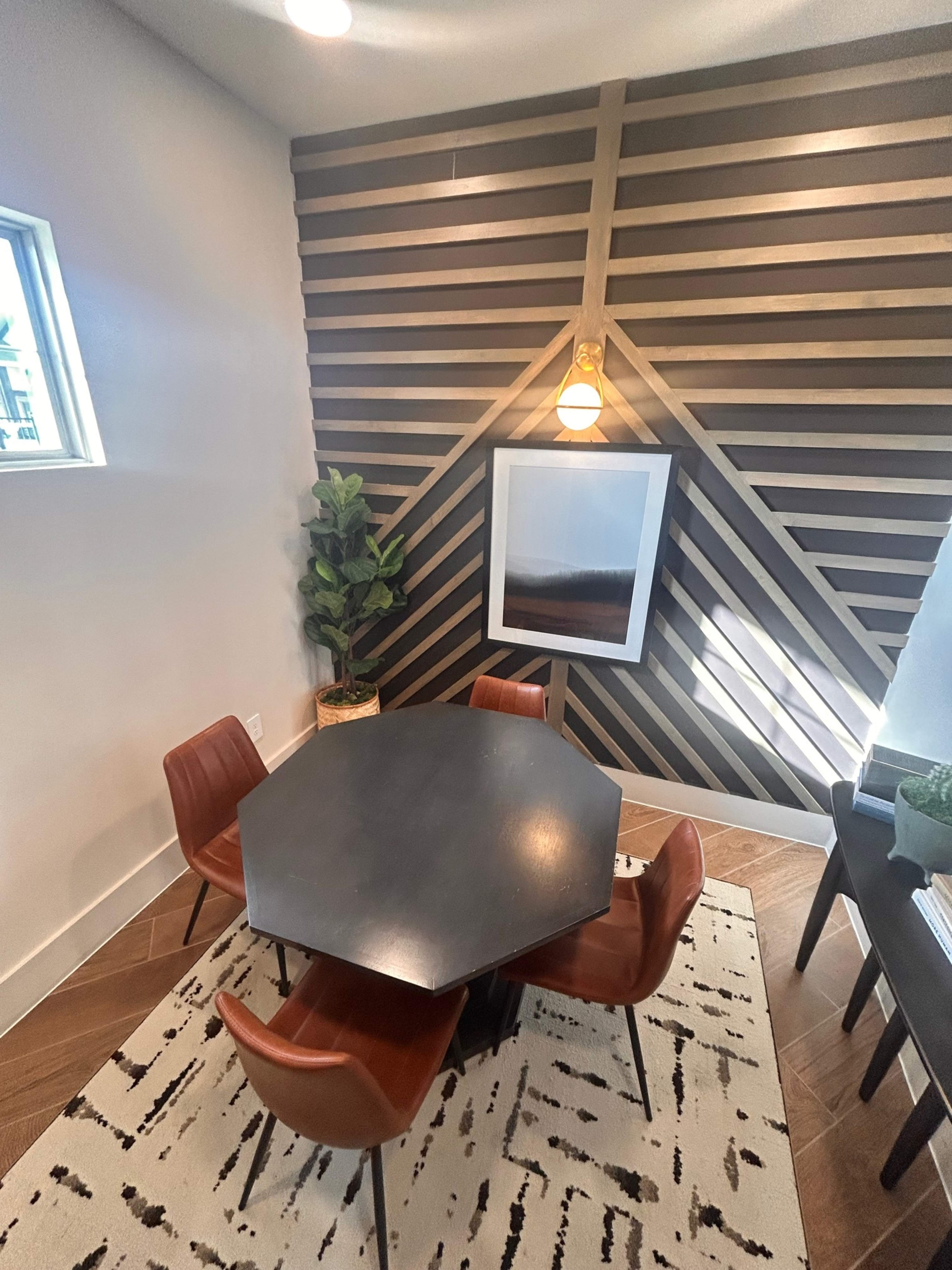 The image shows a modern dining area featuring an octagonal black table surrounded by four rust-colored chairs, with decorative plants and wall art against a wooden slatted wall.