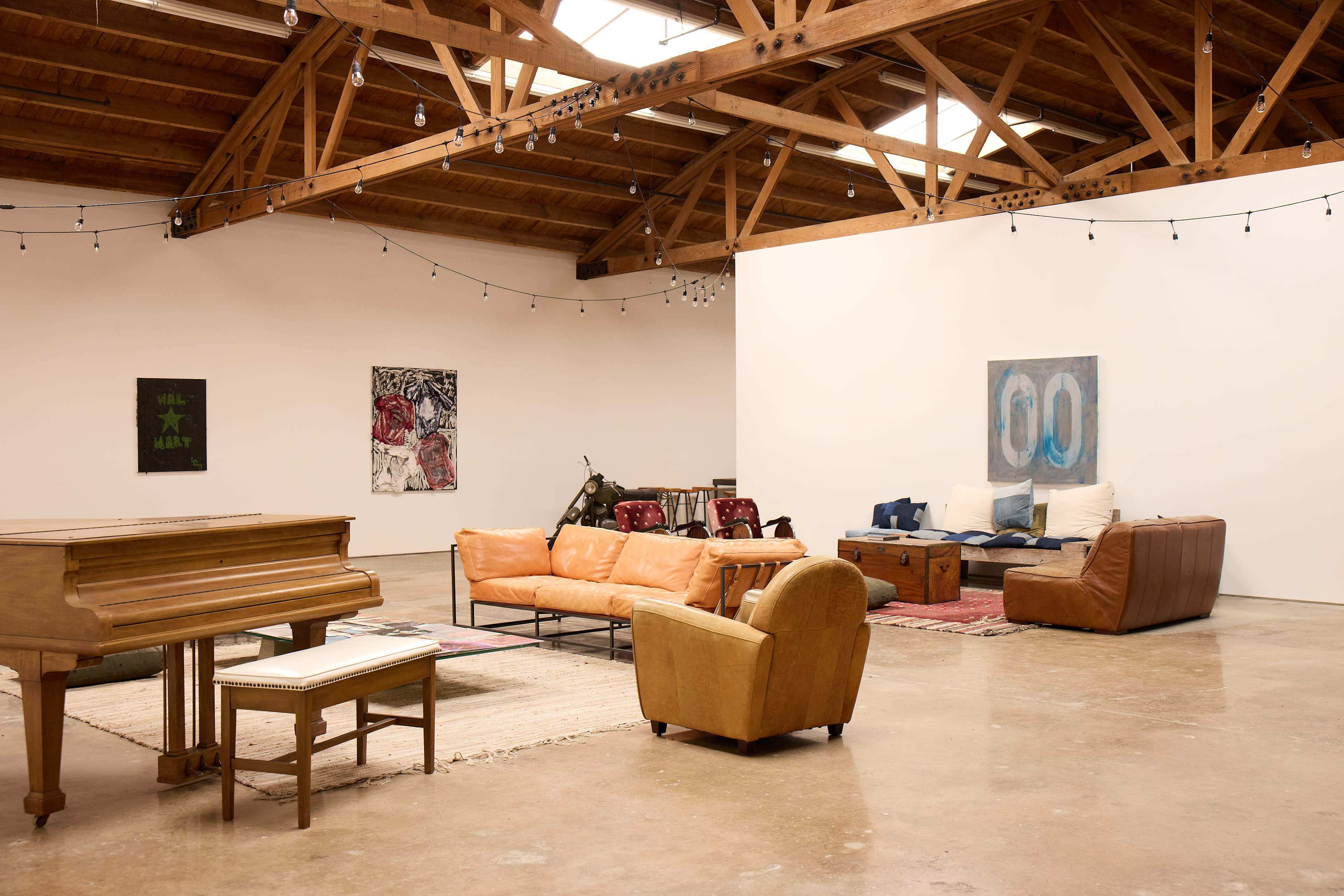 The image shows a spacious room with wooden beams, featuring a piano, various seating arrangements, and artwork on the walls.