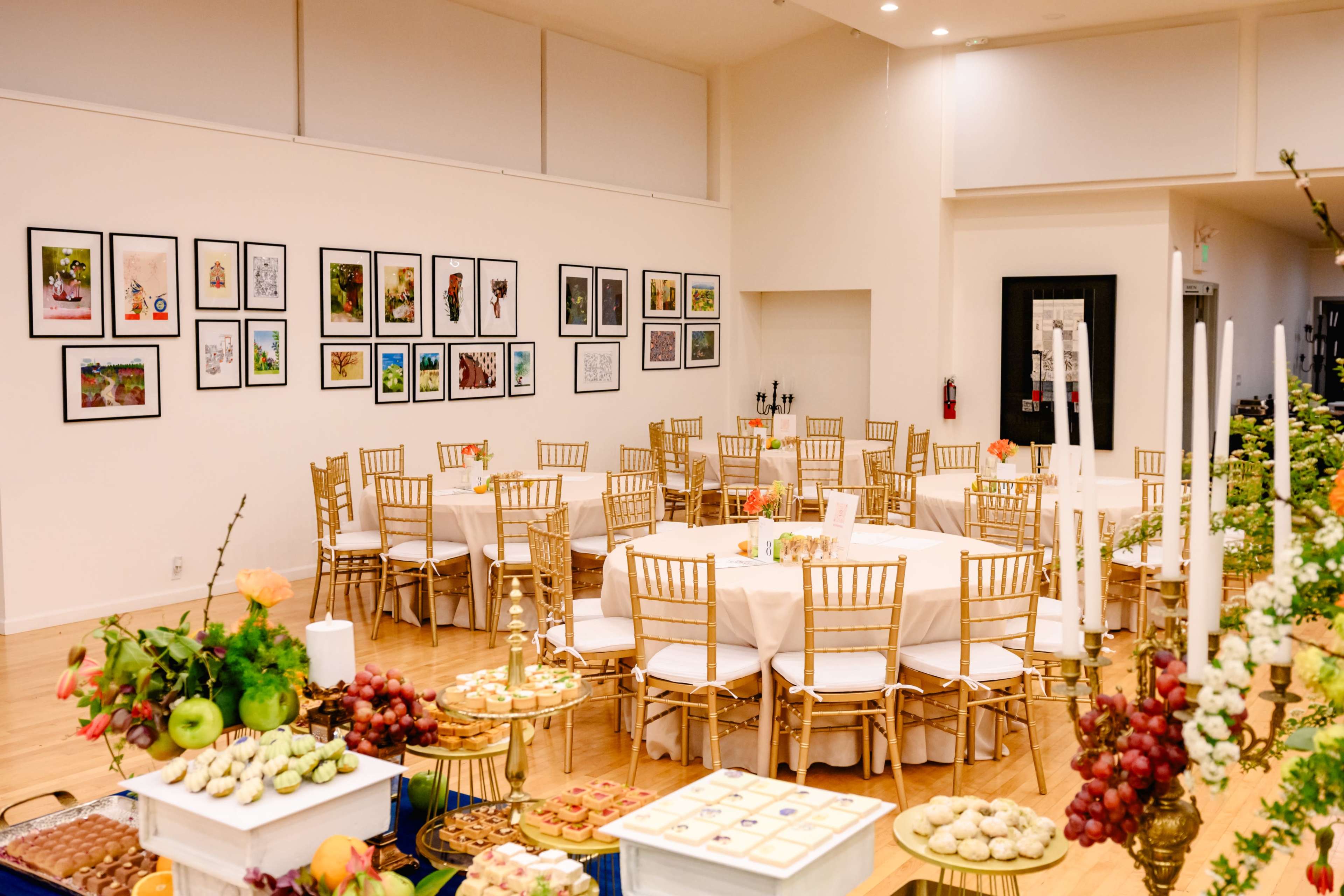 The image shows a dining area set with round tables dressed in white tablecloths and gold chairs, surrounded by framed artworks on the walls and a dessert display featuring various pastries and fruits.
