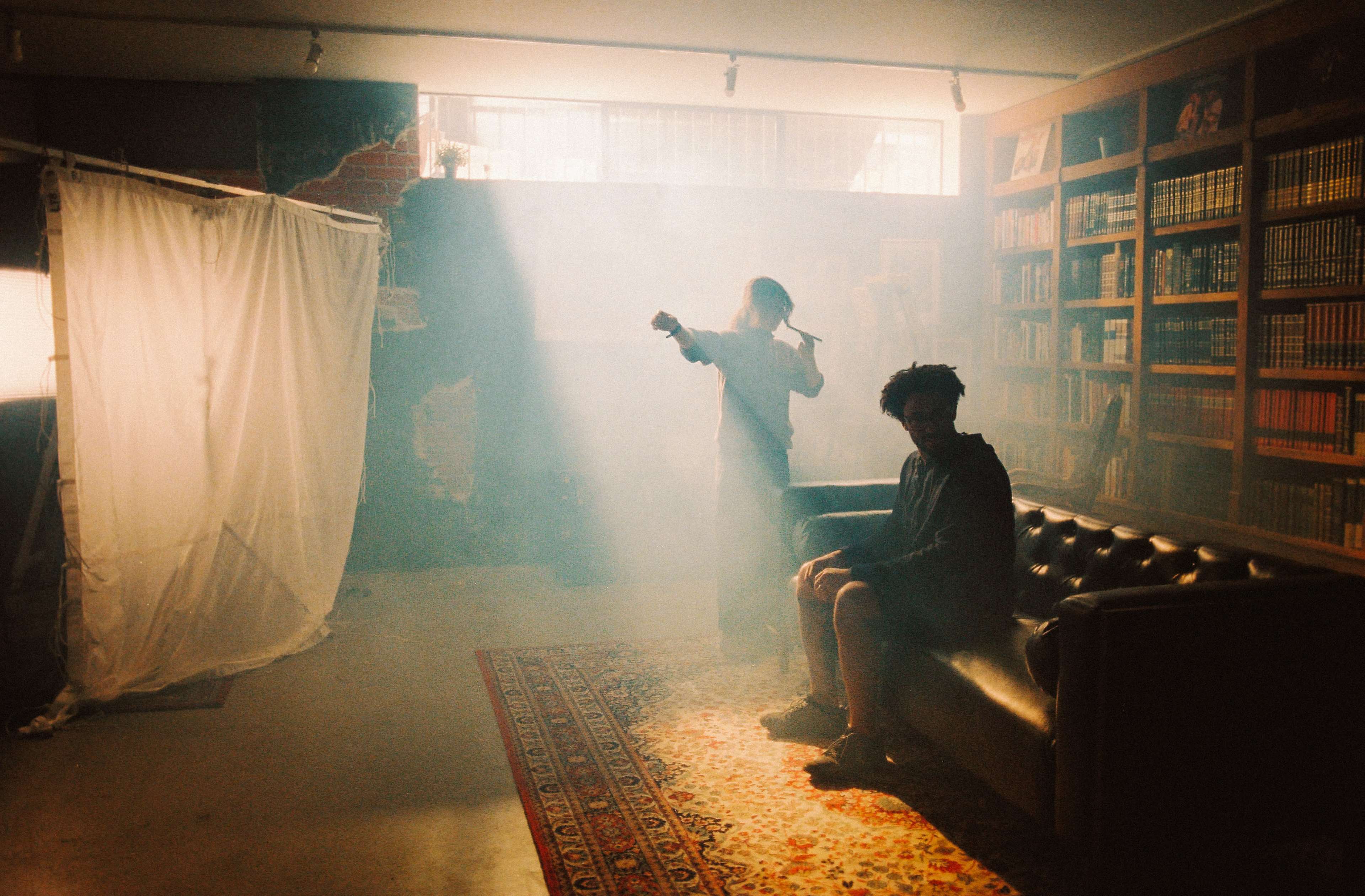 A softly lit room features a person holding a tool in the background while another person sits on a couch in the foreground, with bookshelves lining the walls and a curtain partially drawn.