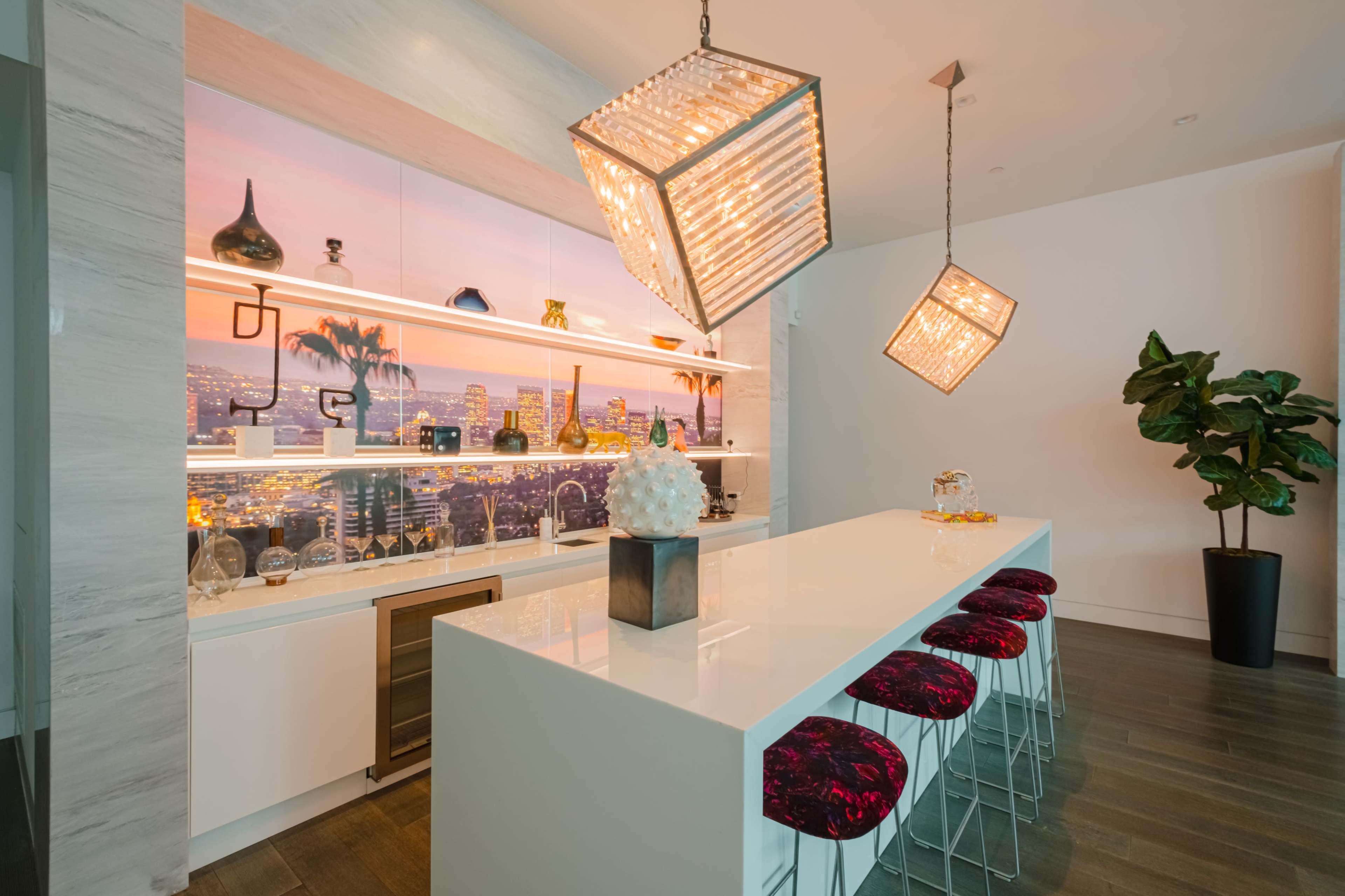 A modern kitchen features a white island with red bar stools, illuminated by two unique pendant lights, against a backdrop of a decorative wall with a city skyline.