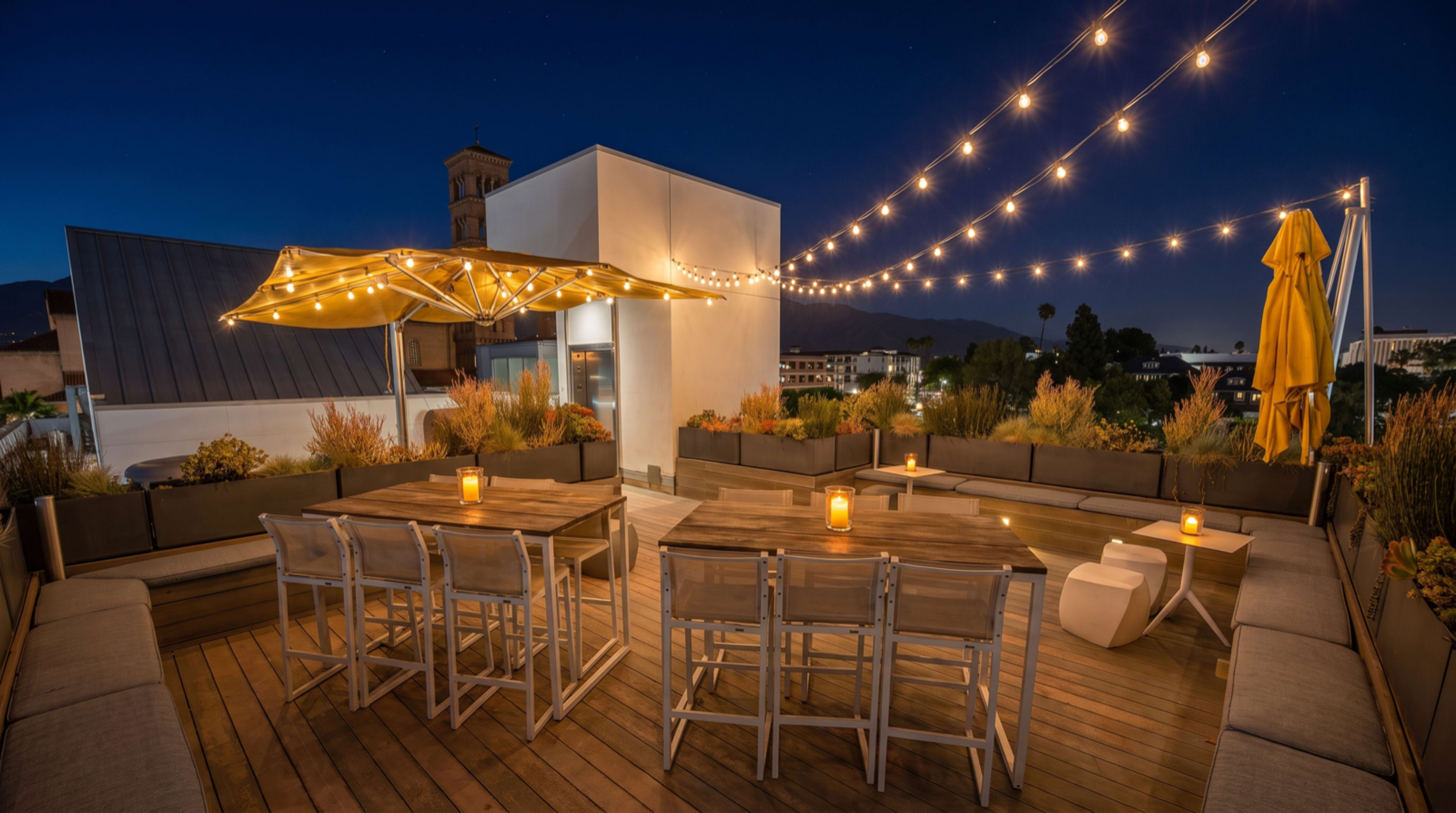 The image shows a rooftop terrace illuminated with string lights and featuring a dining table, chairs, and an umbrella.