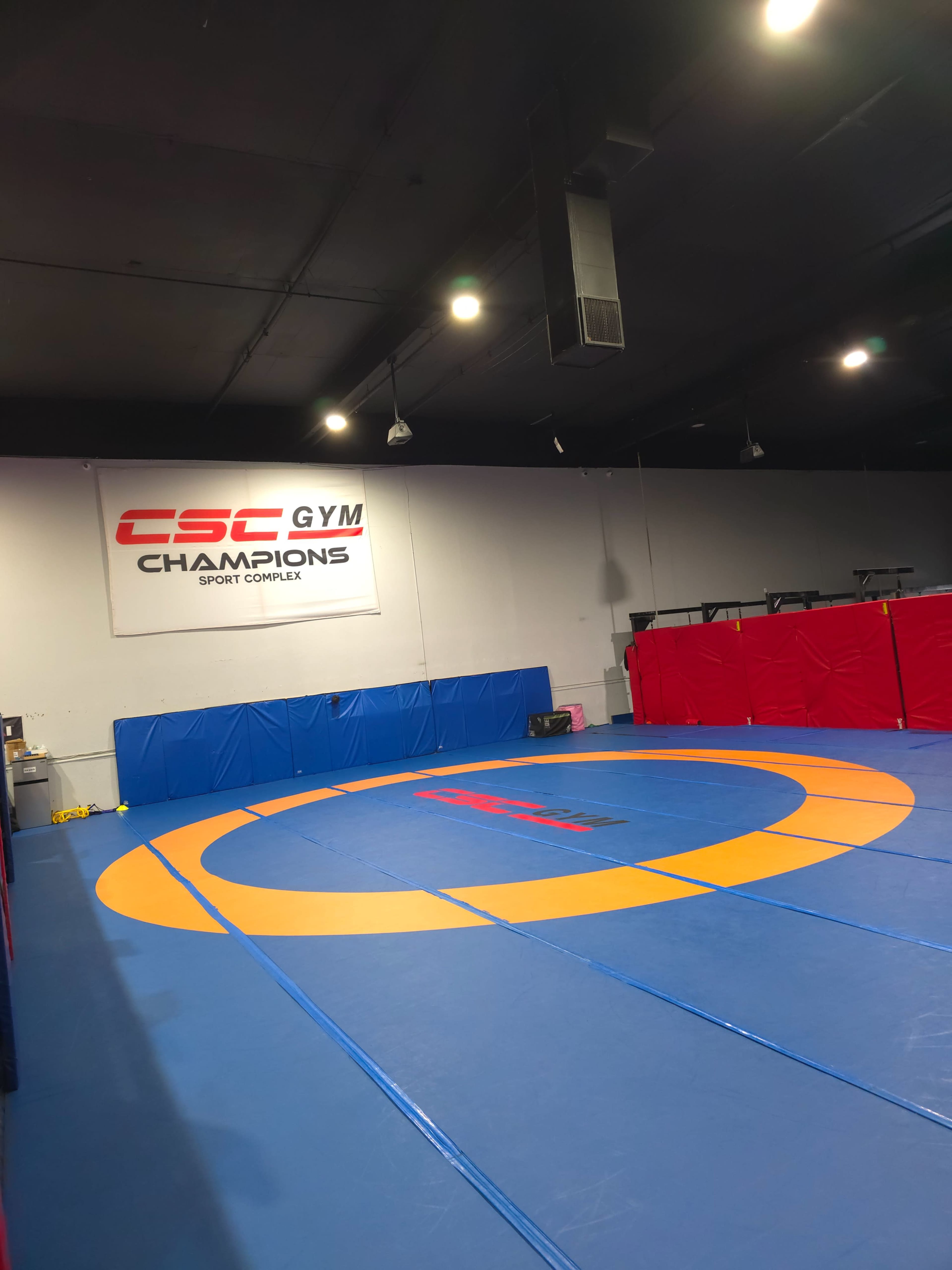 The image shows an indoor wrestling or training facility with a blue and yellow wrestling mat and padded red walls.
