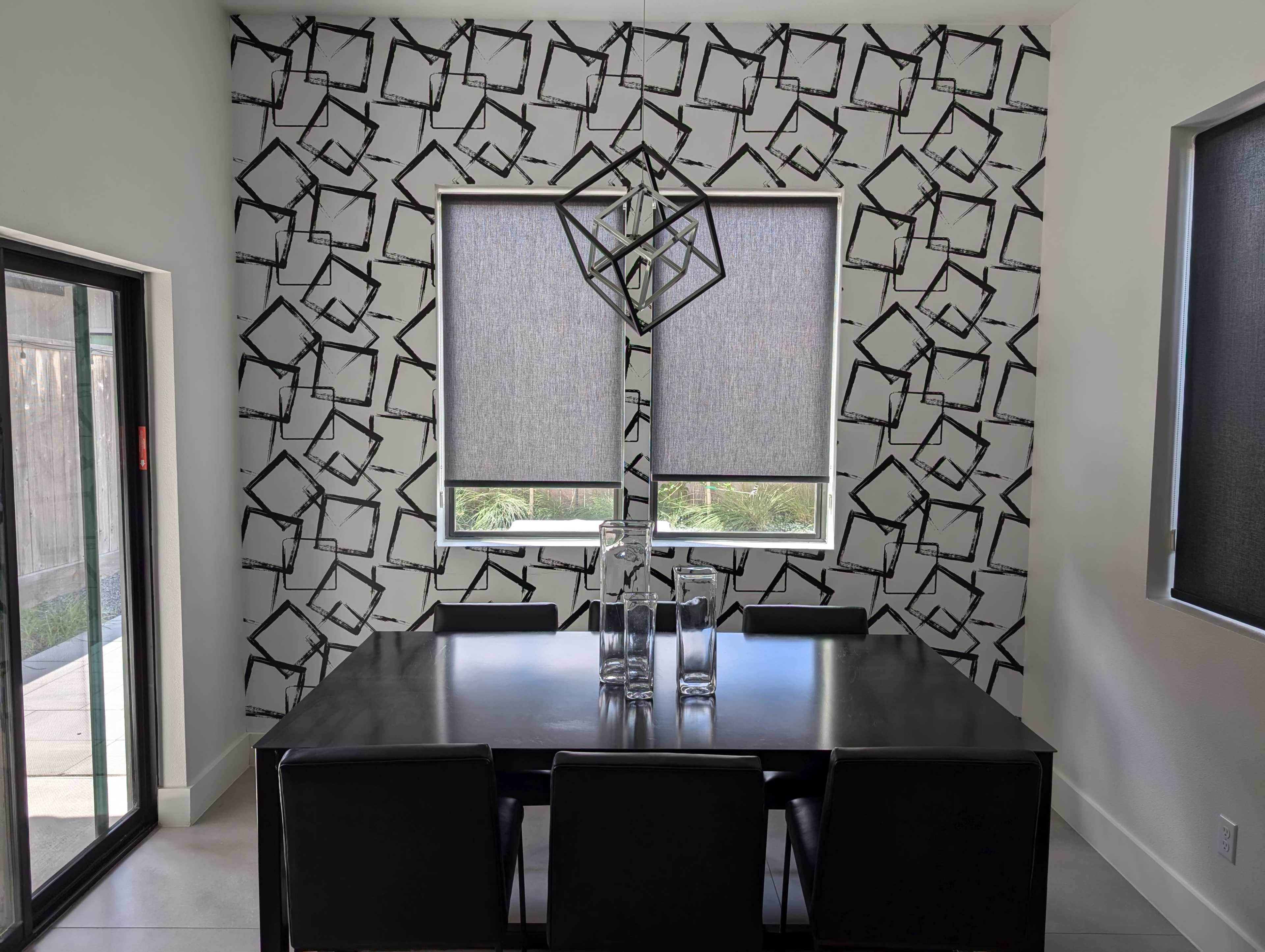 A dining area features a black and white patterned wall, a rectangular table with black chairs, and two large windows with roller shades.
