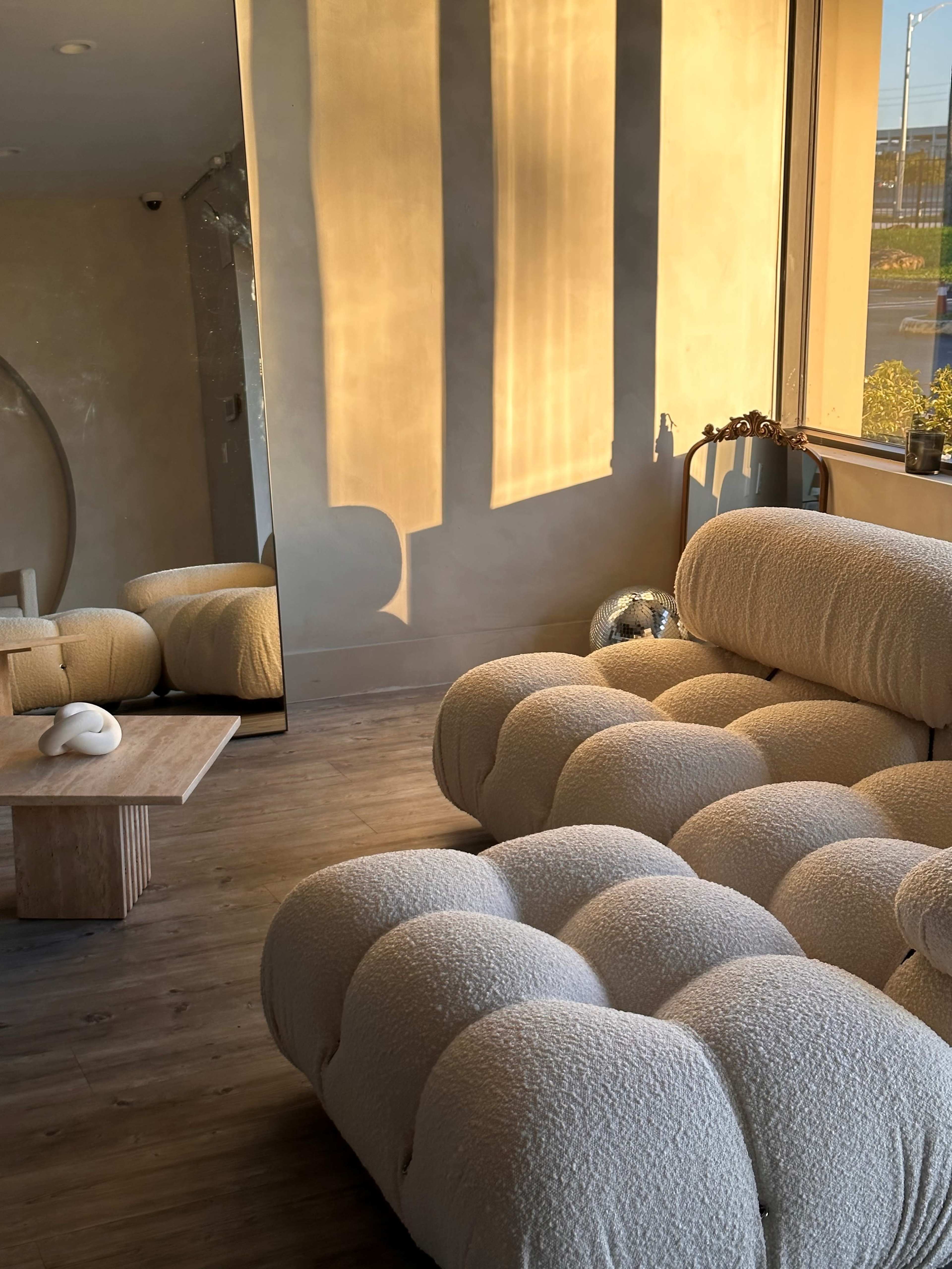 A plush, textured sofa sits next to a large window, reflecting warm sunlight onto a minimalist wooden coffee table and a nearby mirror.