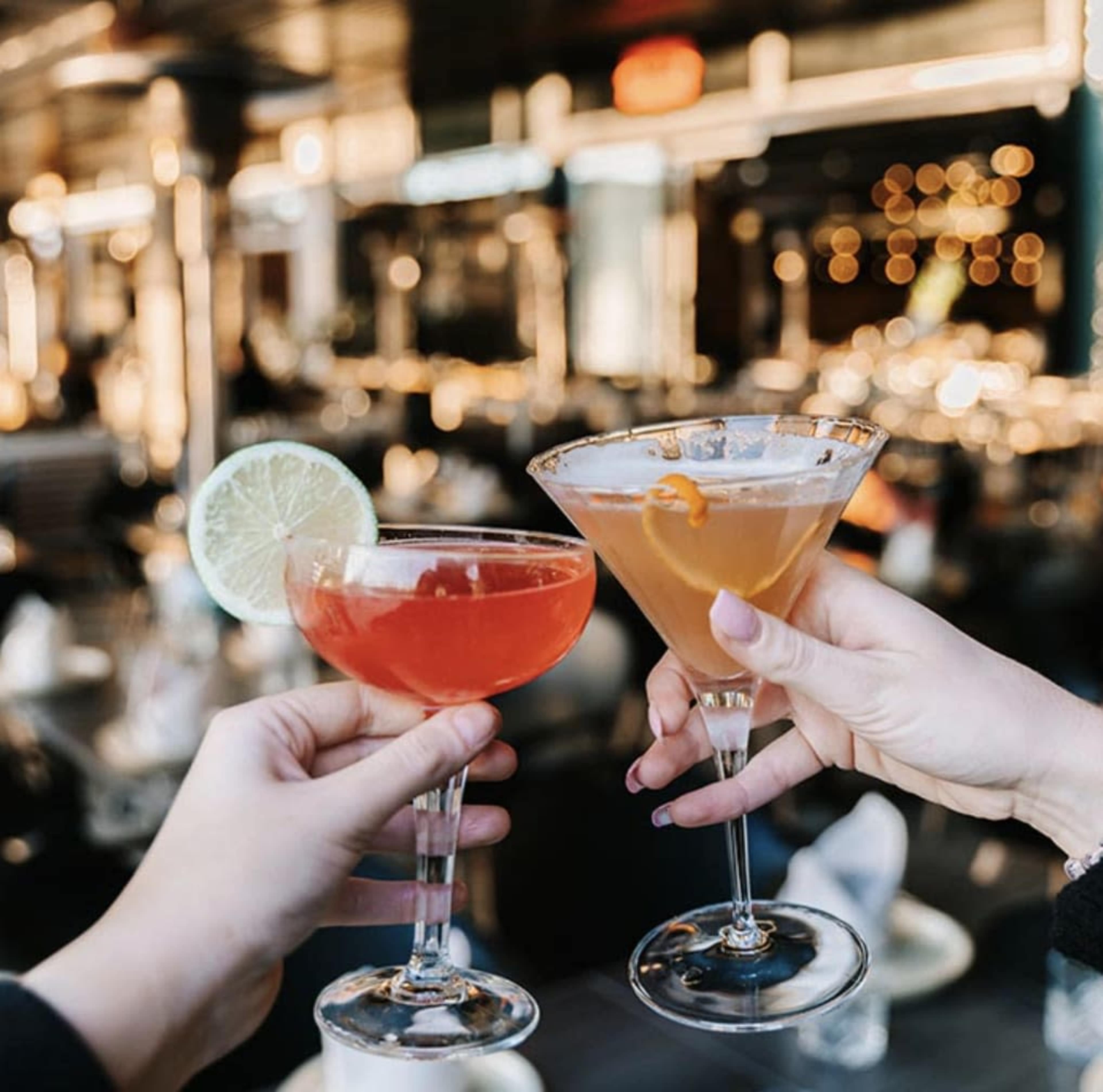 Two hands hold cocktail glasses, one with a lime slice and the other garnished with an orange twist, against a blurred bar background.