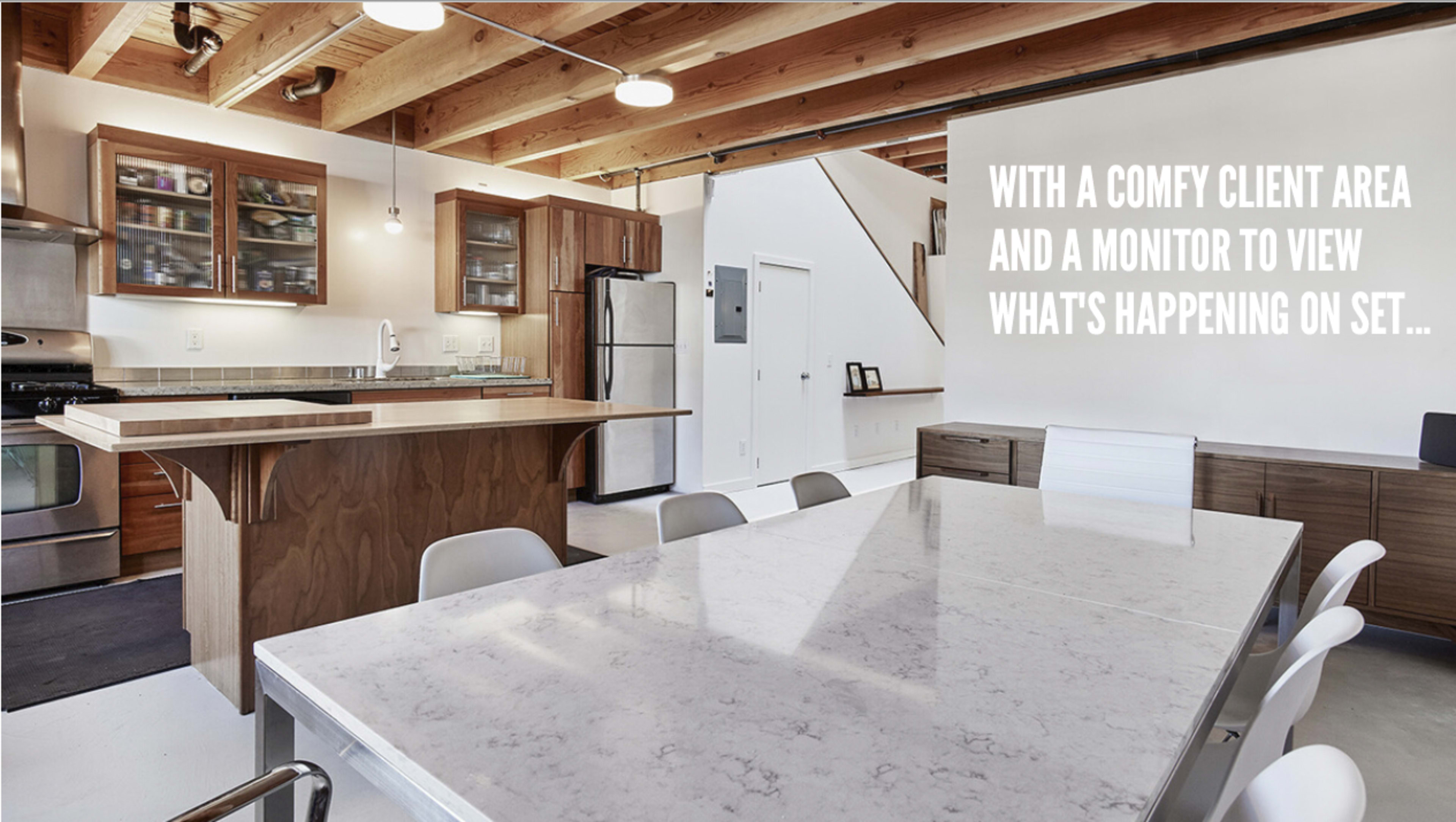 A modern kitchen and dining area with a large marble table, sleek cabinetry, and a monitor mounted on the wall for viewing activities on set.