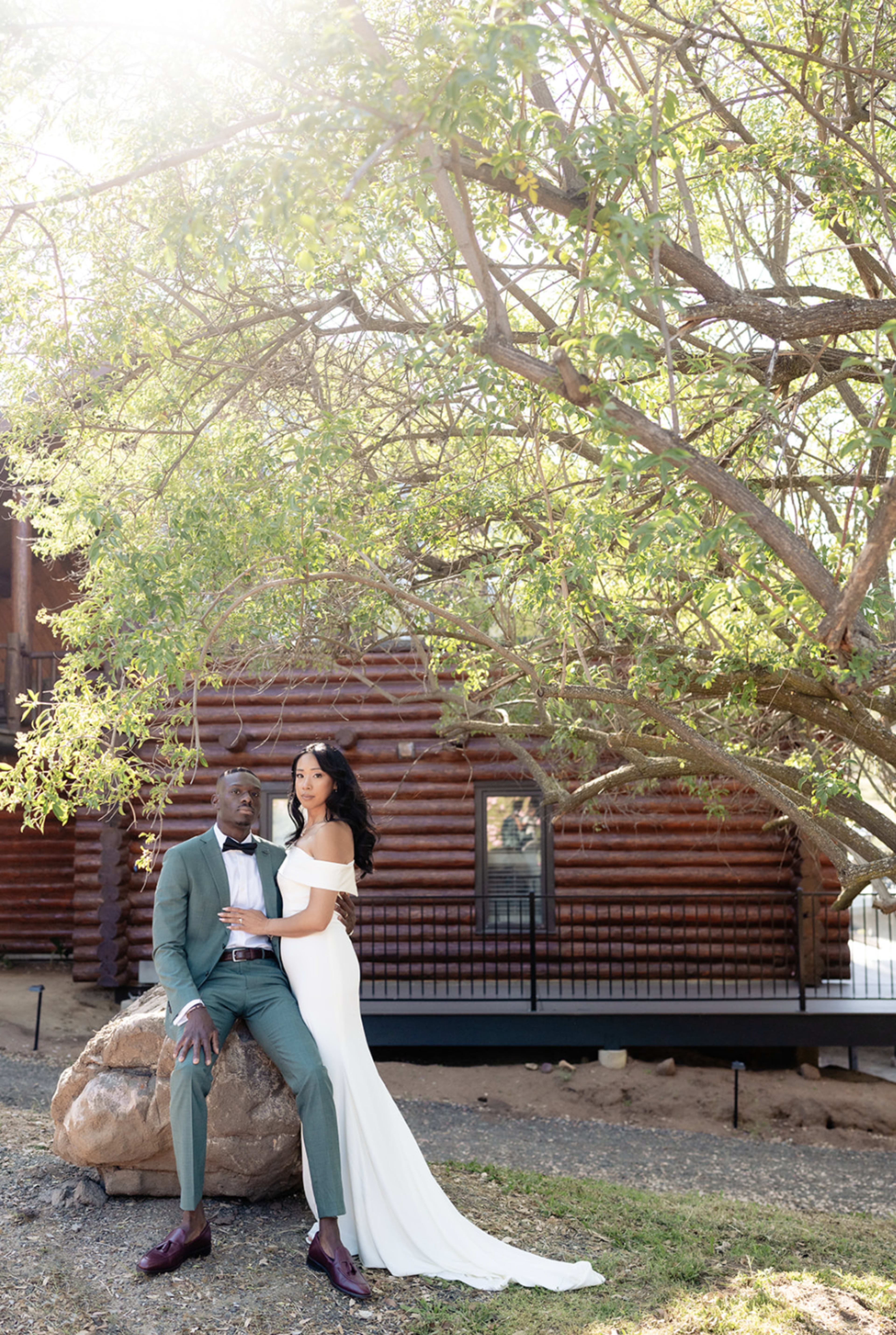 A couple poses together under a tree near a log cabin.