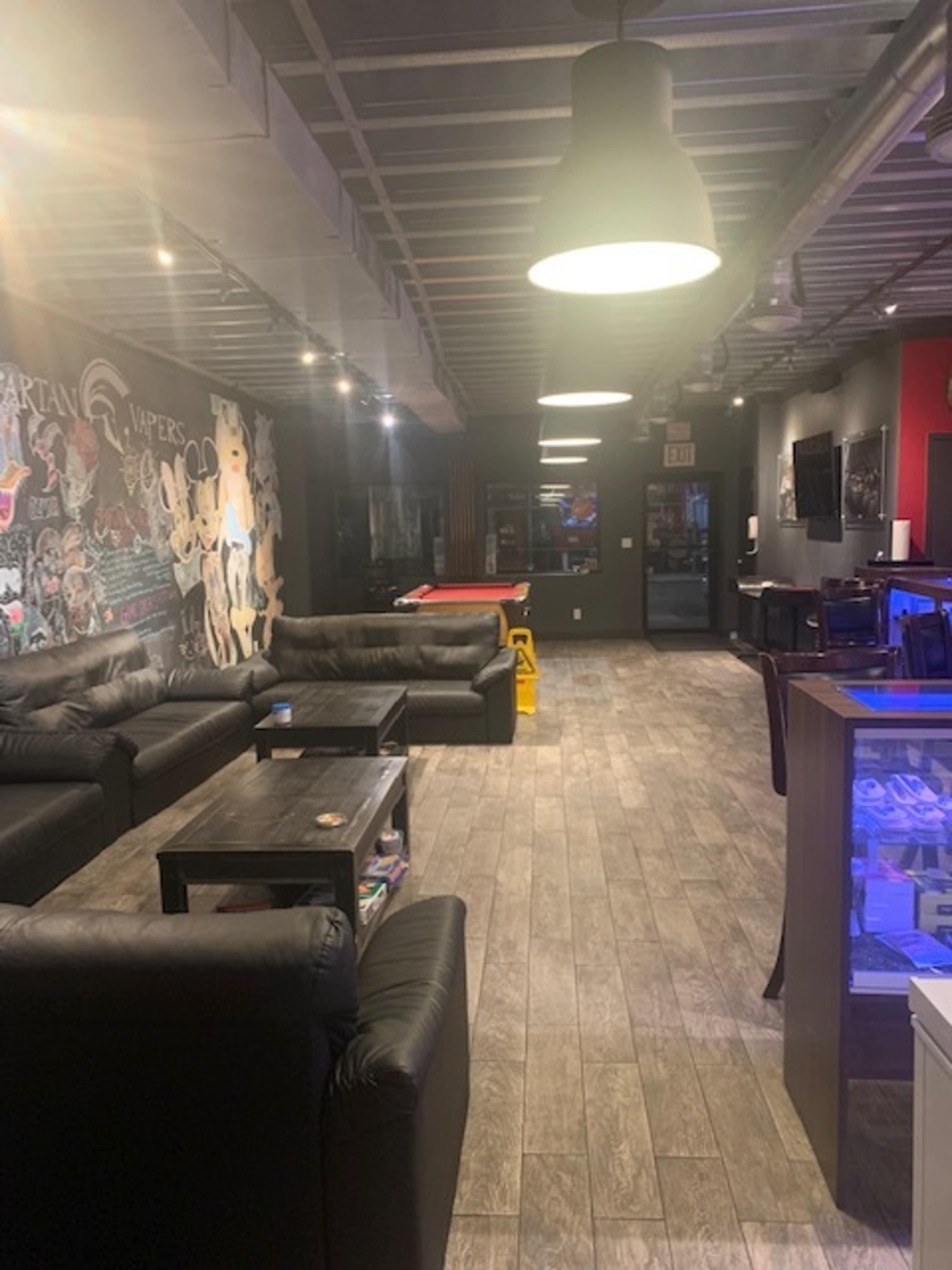A dimly lit lounge area with black leather sofas, wooden tables, and a mural on the wall, with a pool table at the far end.
