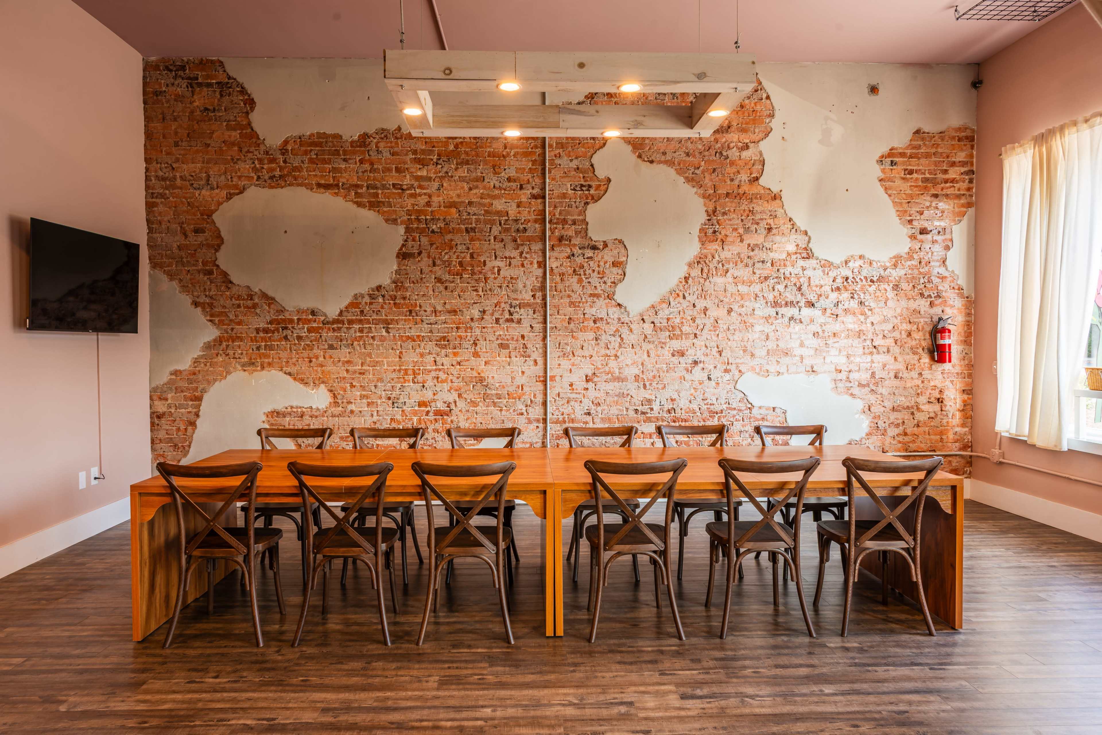 The image shows a long wooden dining table with chairs arranged around it, set against an exposed brick wall that exhibits a distressed look.